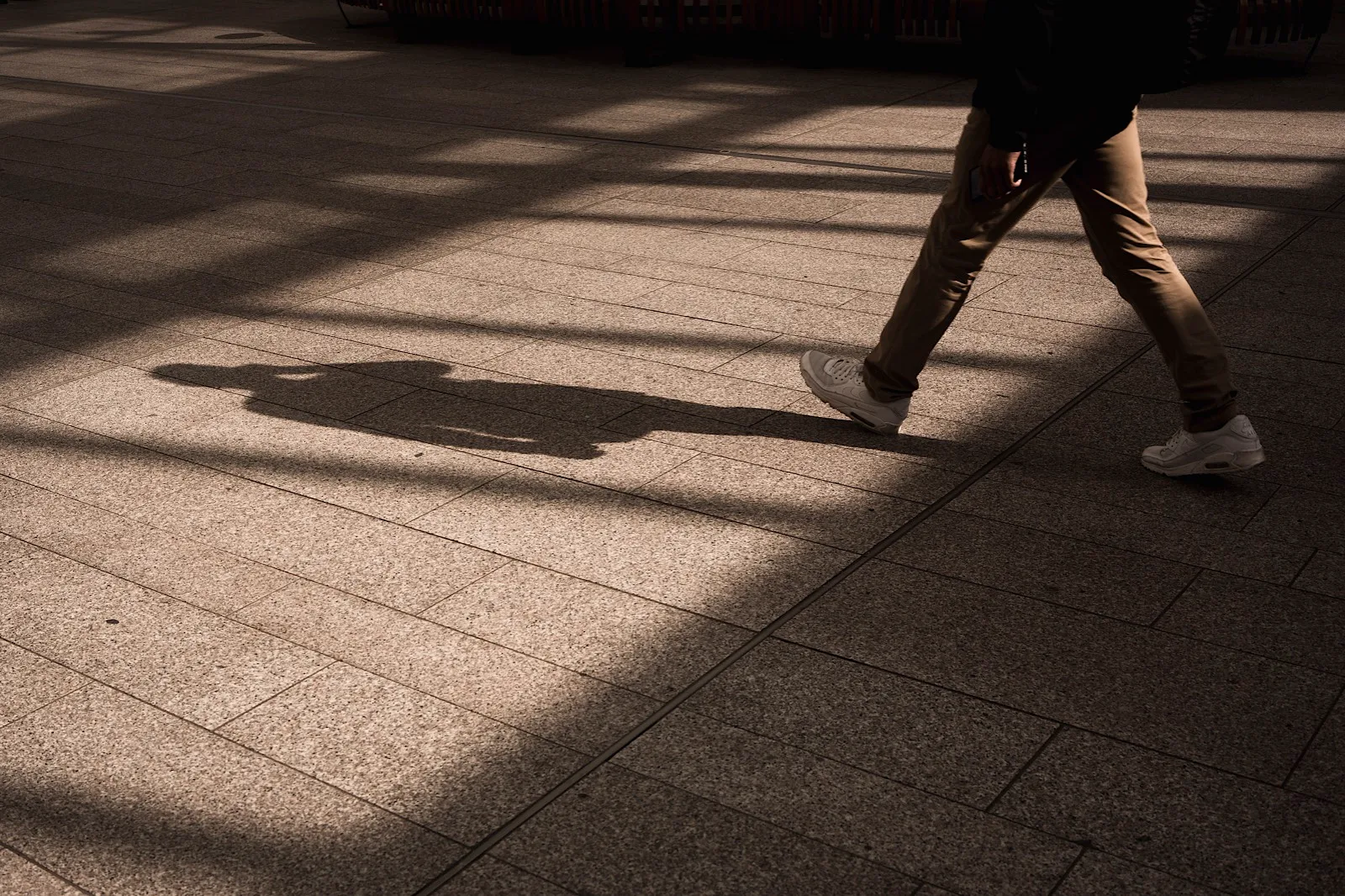 Person walking on a sunlit pavement, casting a long shadow. Only their legs are visible, wearing tan pants and white sneakers.