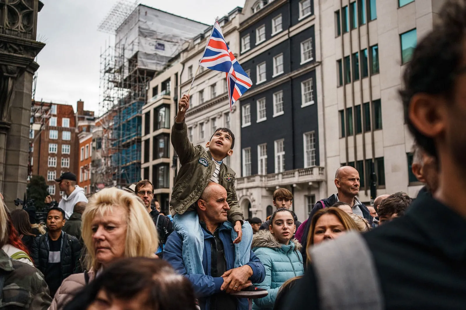 Young boy on man's shoulders amongst the crowds during Queen Elizabeth's funeral waving a Union Jack