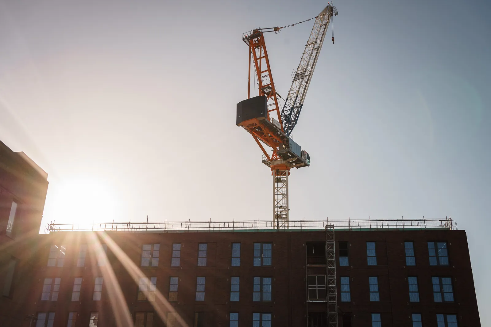 A construction crane towers over a brick building under a clear sky, with the sun shining bright.