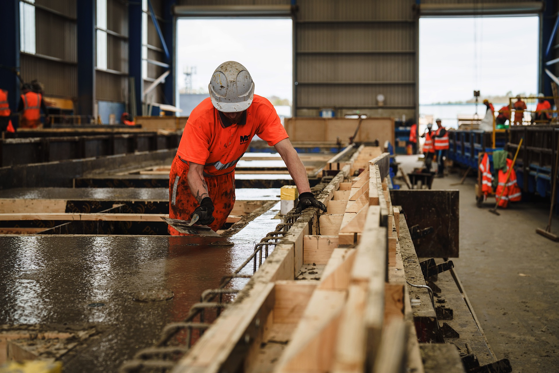 Worker in a construction site wearing a hard hat and orange vest smooths concrete in a large industrial building. Other workers are visible in the background.