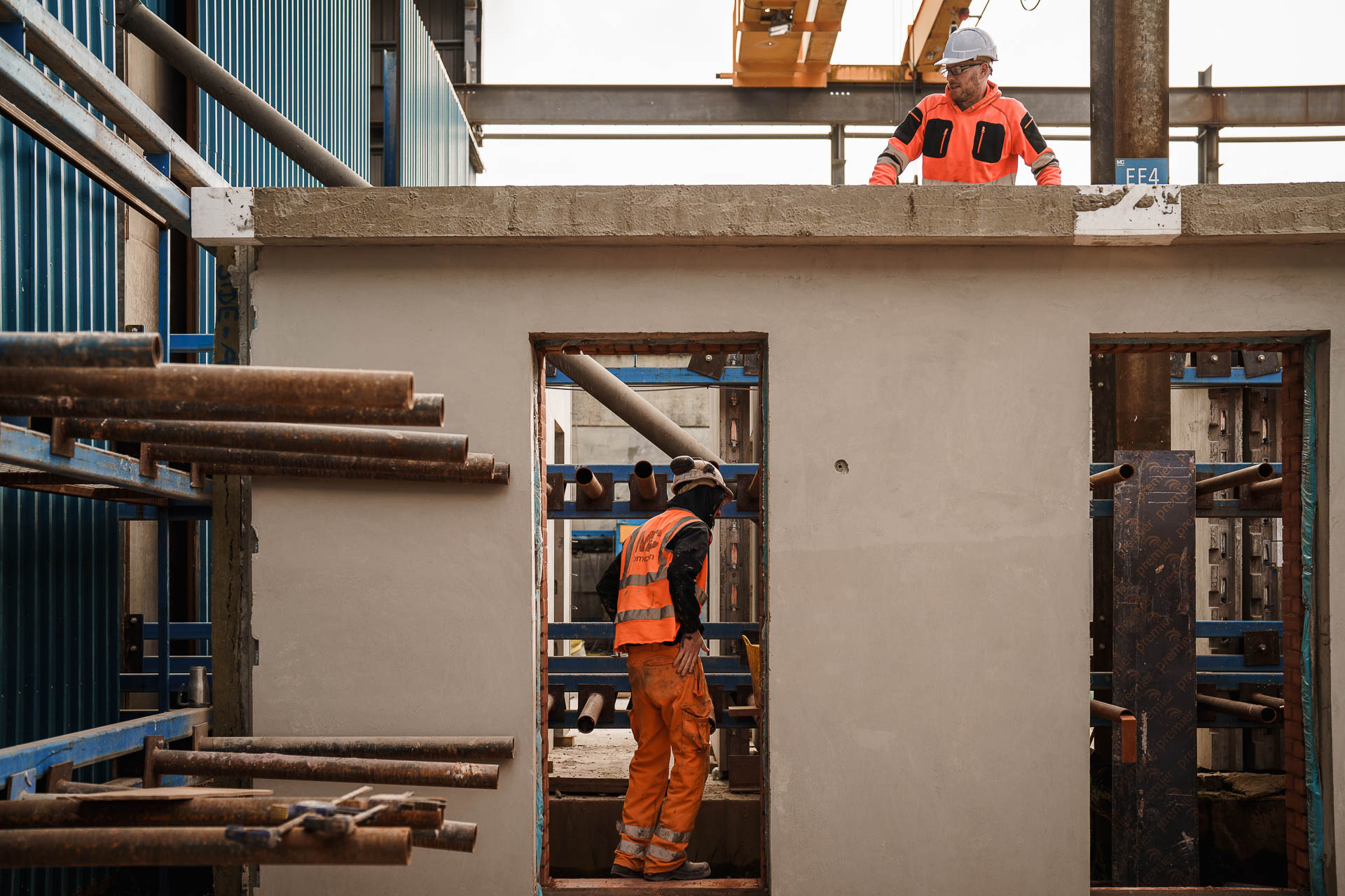 Two construction workers in safety gear work on a building site, one on a concrete structure and the other standing below in an opening, surrounded by scaffolding and building materials.