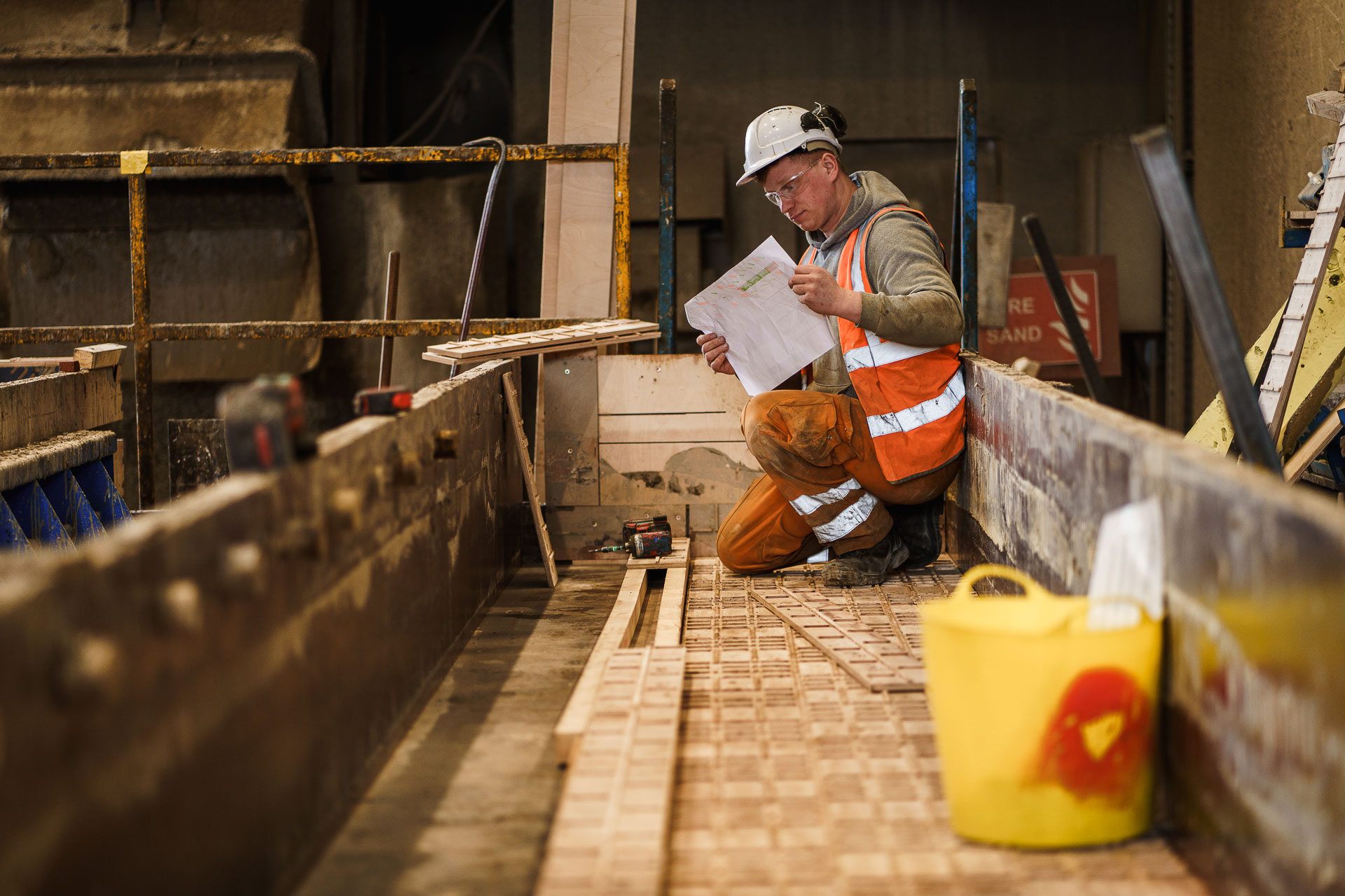 Construction worker in safety gear kneels on a wooden surface, examining documents at a building site.