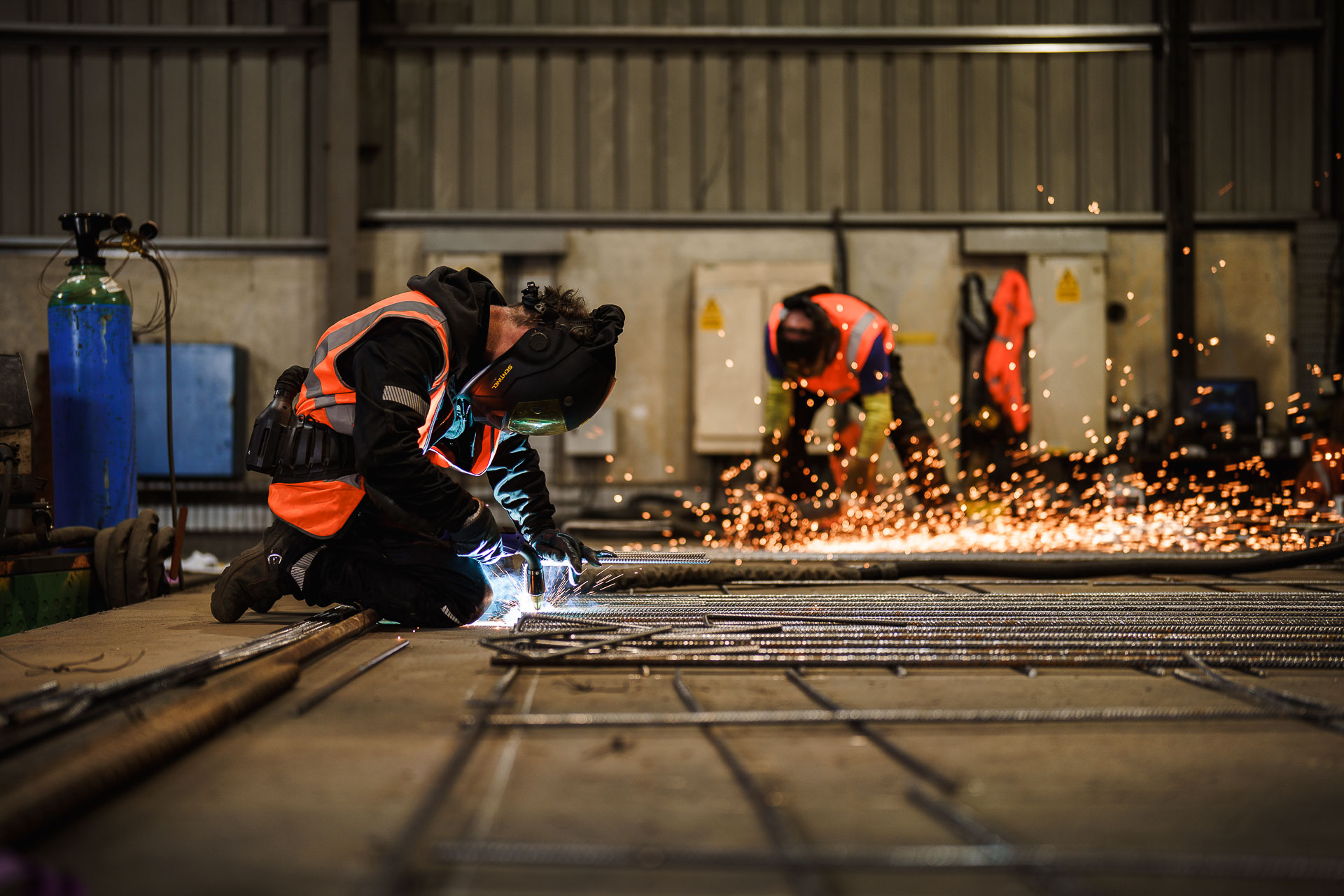 Two workers in safety gear are welding and grinding metal in an industrial workshop, with sparks flying from their tools.