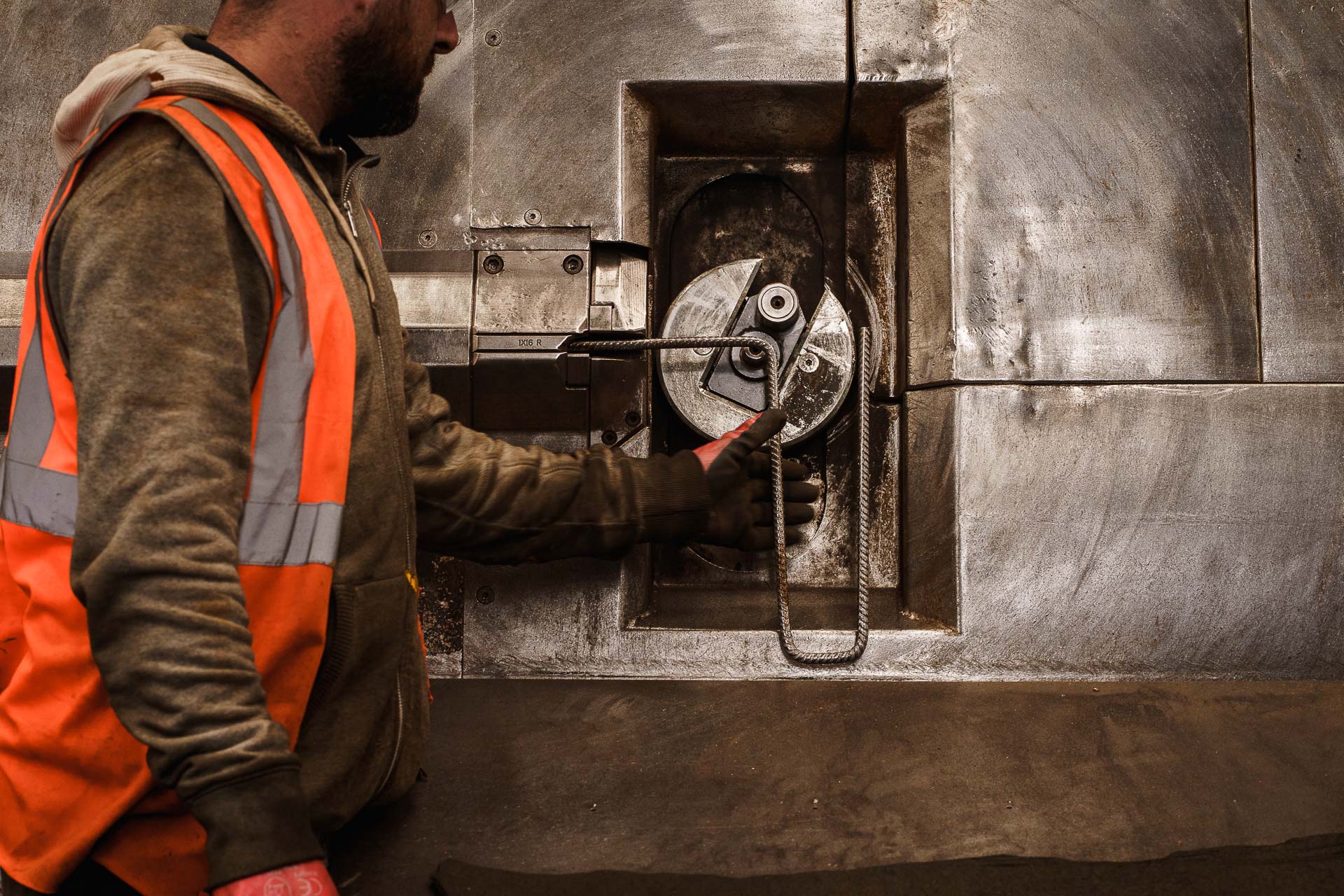 Person in high-visibility vest operates industrial machinery with manual controls, surrounded by metallic surfaces.