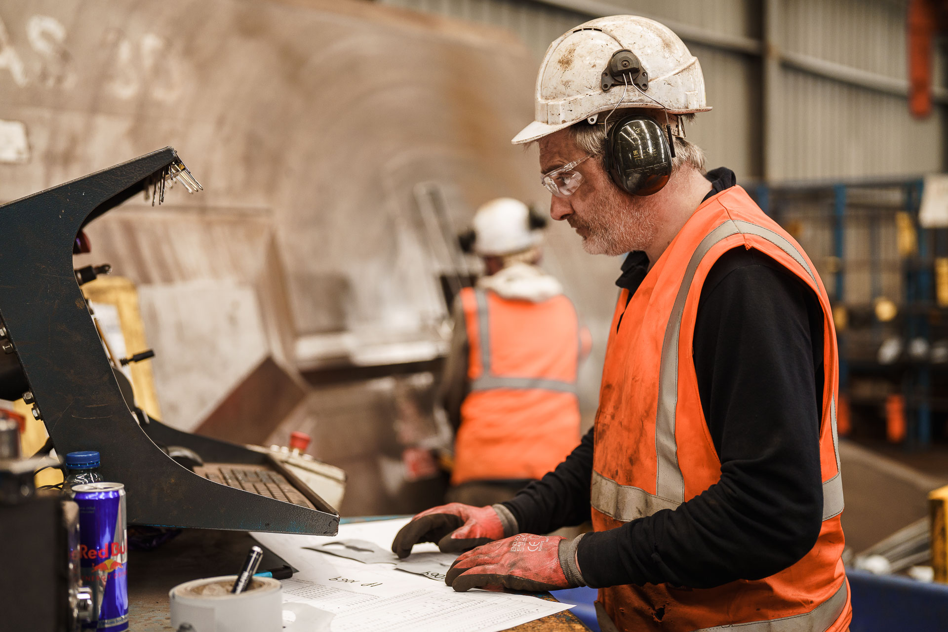 A worker wearing a helmet, ear protection, and high-visibility vest operates machinery in an industrial setting, with another worker visible in the background.