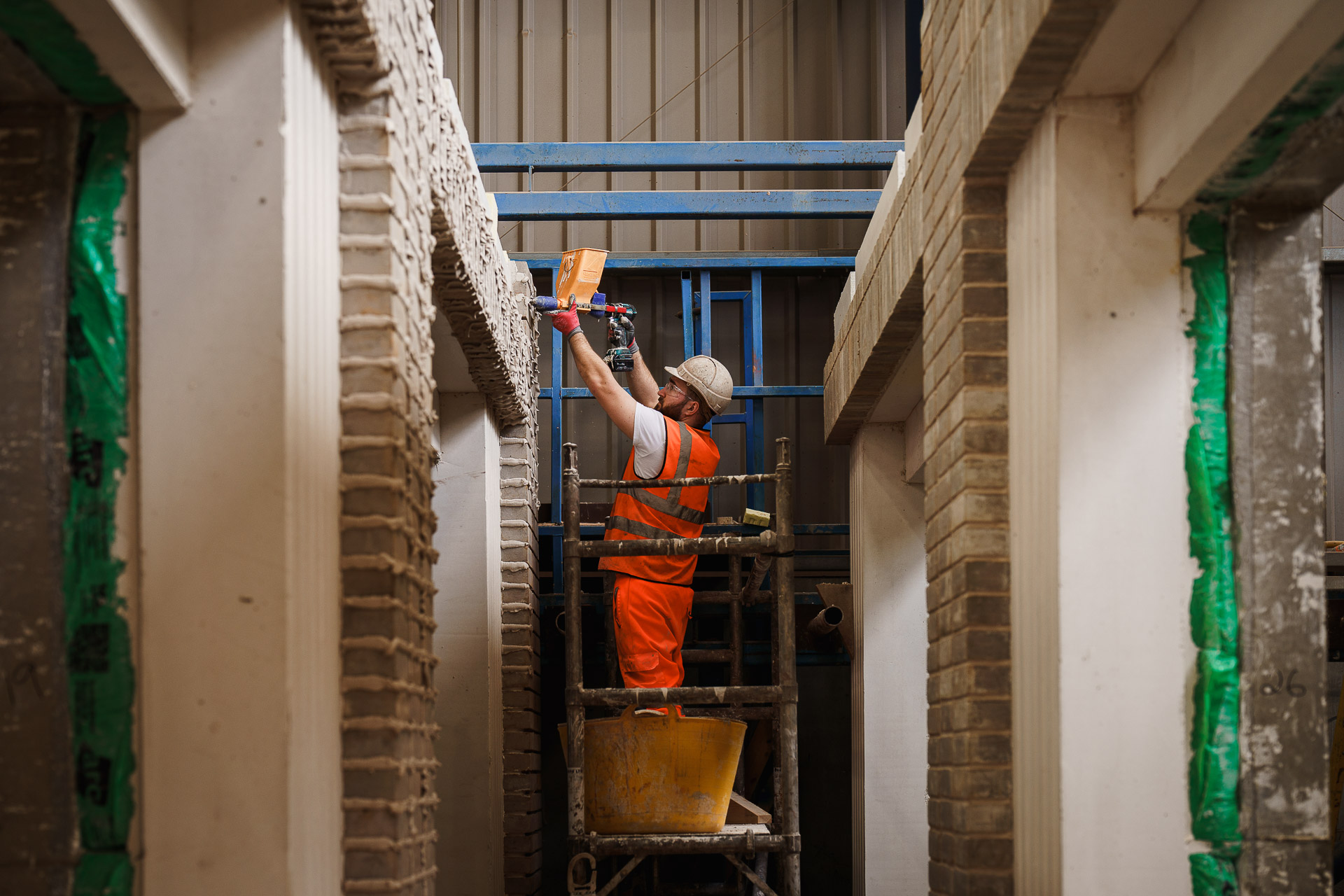 Construction worker in an orange vest and helmet standing on scaffolding, applying mortar to a brick wall with a trowel in an interior space.