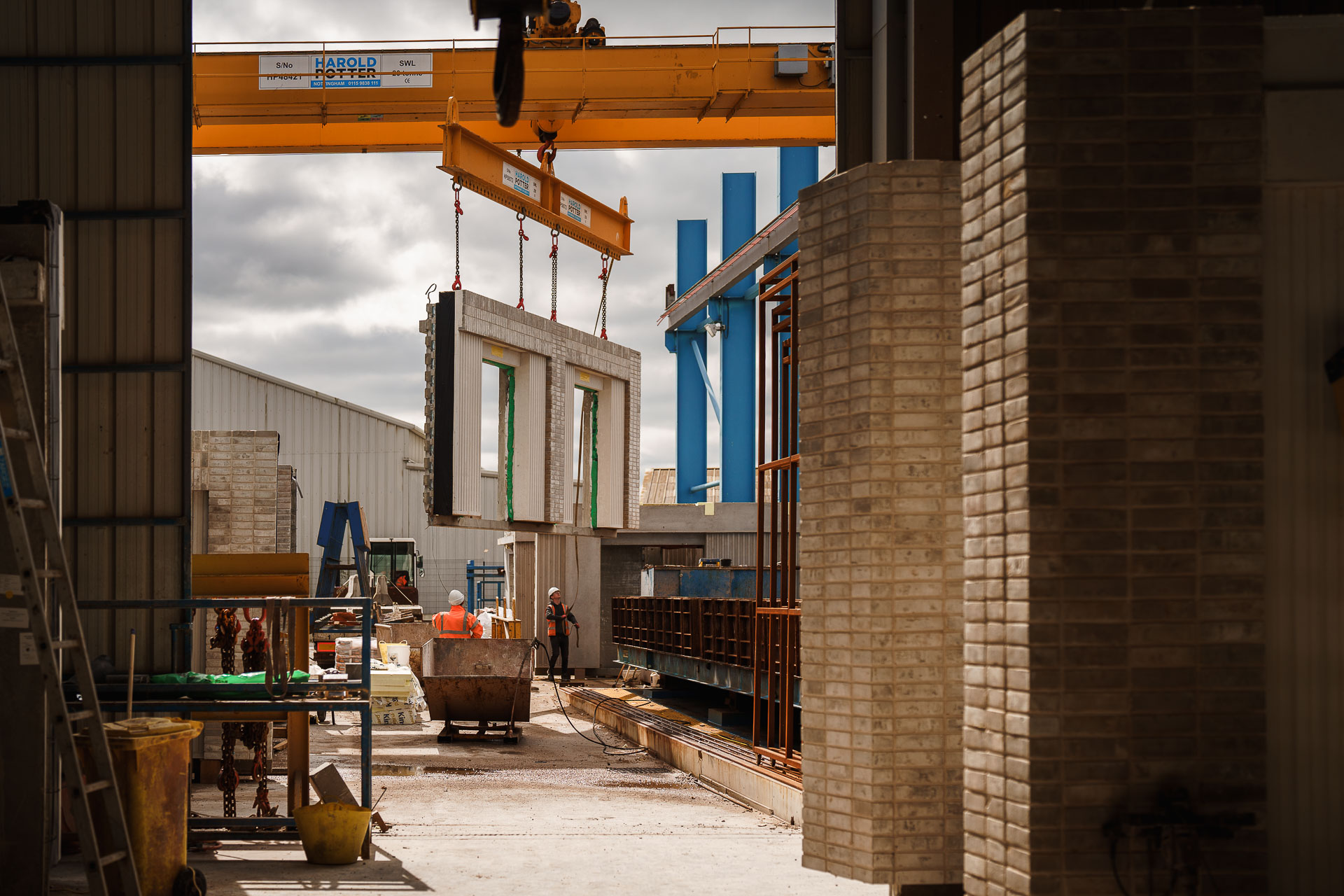 A construction site with a crane lifting a concrete wall panel. Workers and various construction materials are visible, with buildings and equipment in the background.