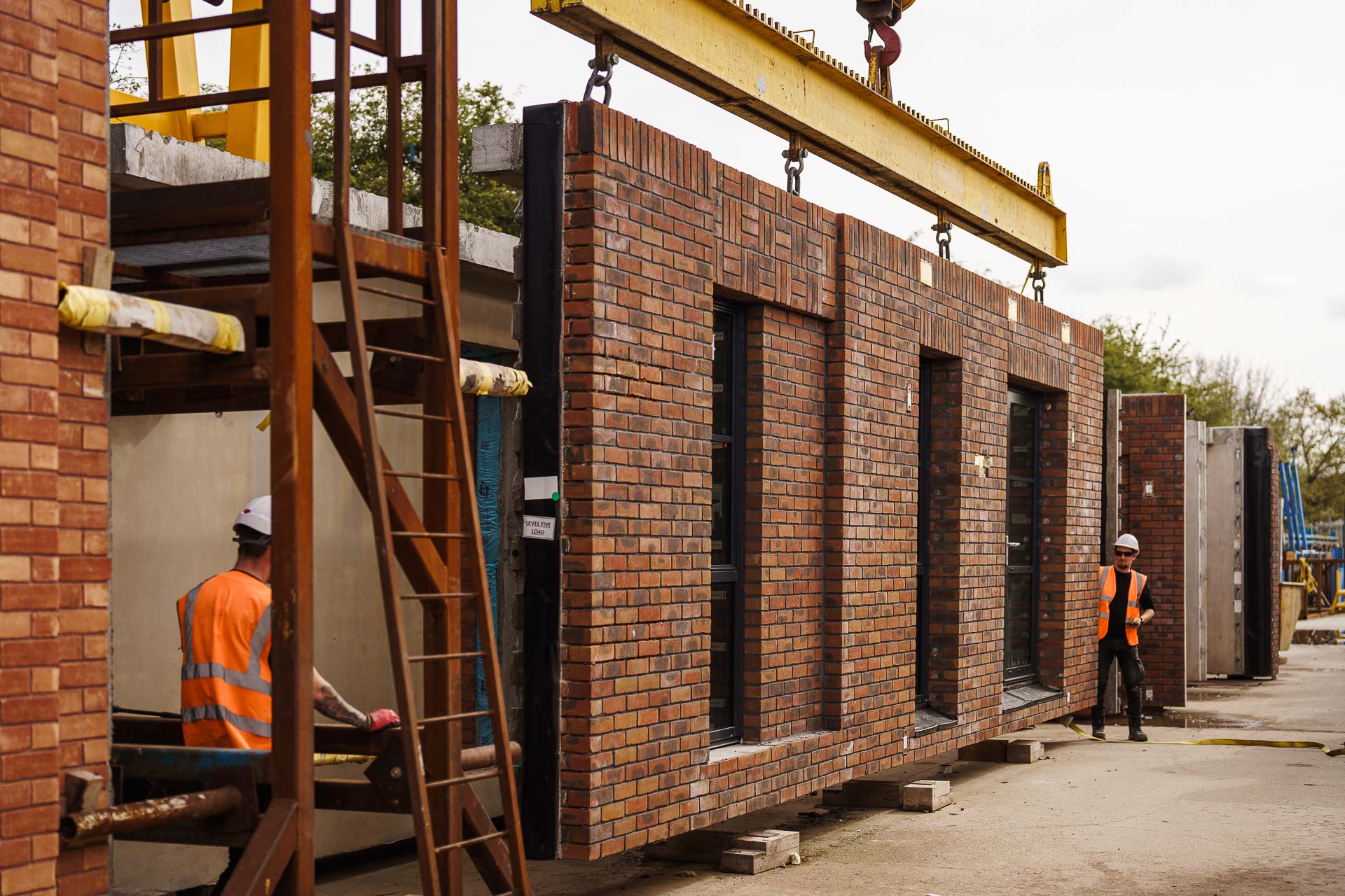 Construction site with workers installing large prefabricated brick walls using a crane. Workers wear safety vests and helmets.