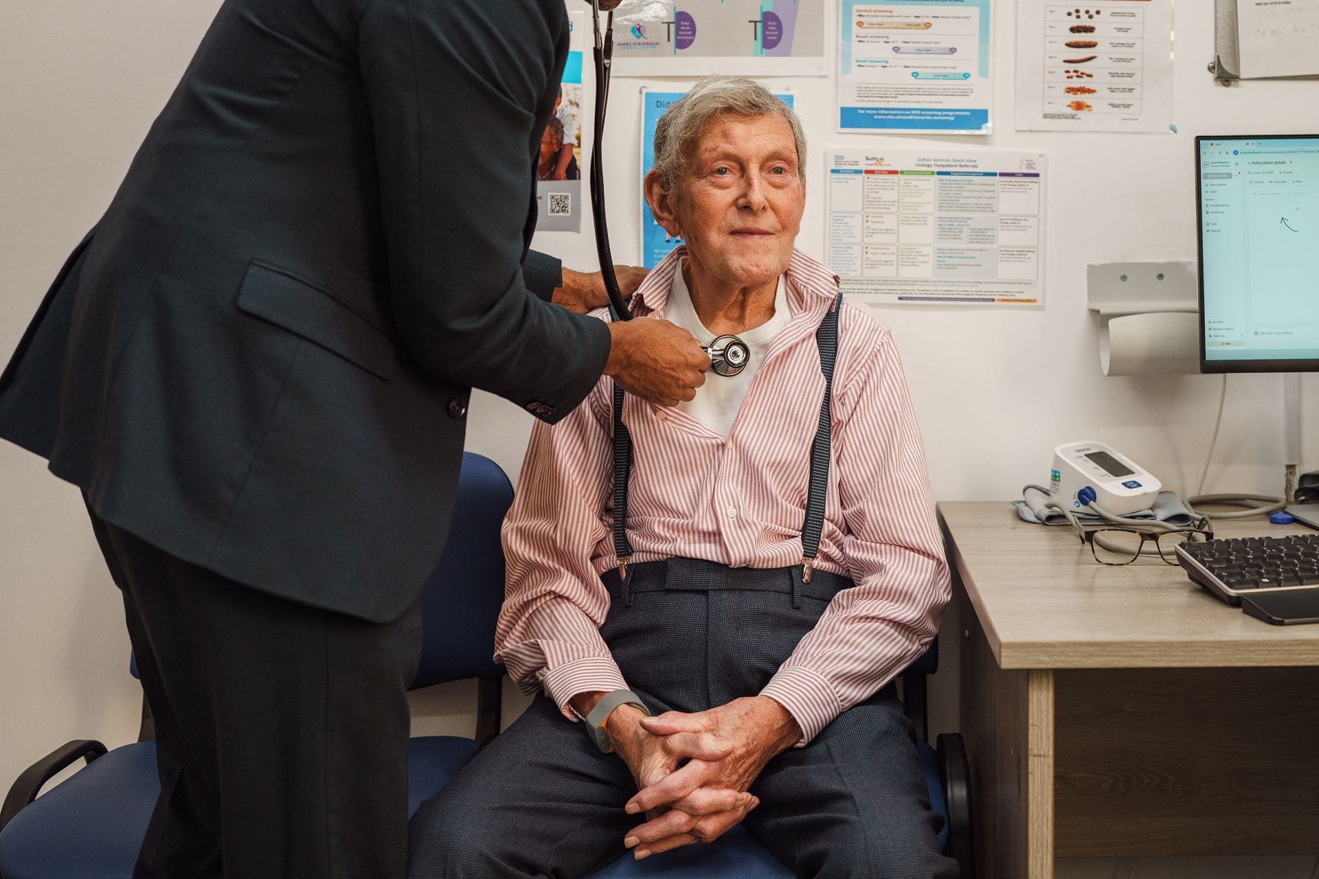 A doctor uses a stethoscope to examine an older man sitting in a medical office. The man wears a striped shirt; medical equipment and posters are visible in the background.