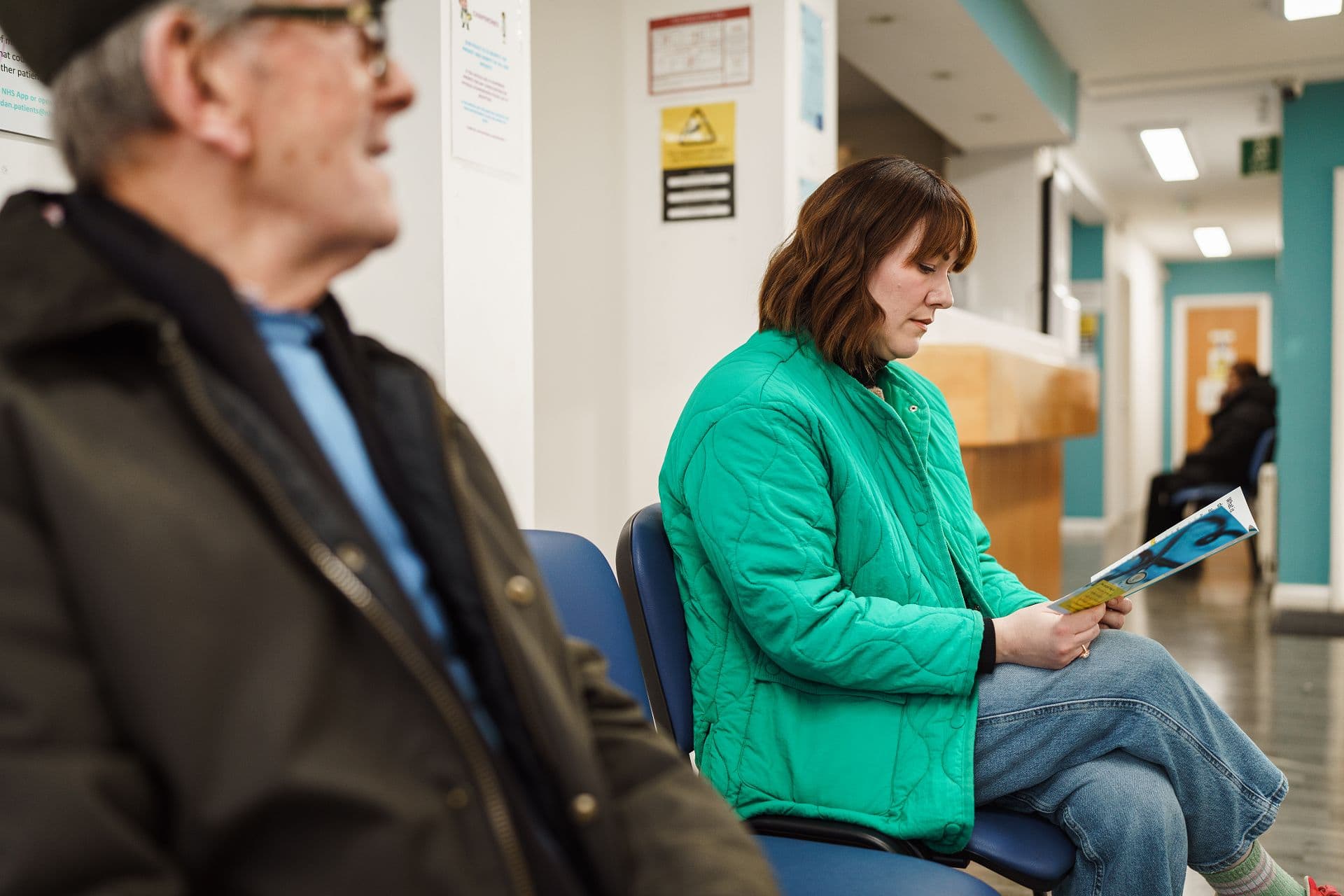 A woman in a green jacket reads a magazine while sitting in a waiting area, with an older man seated nearby.