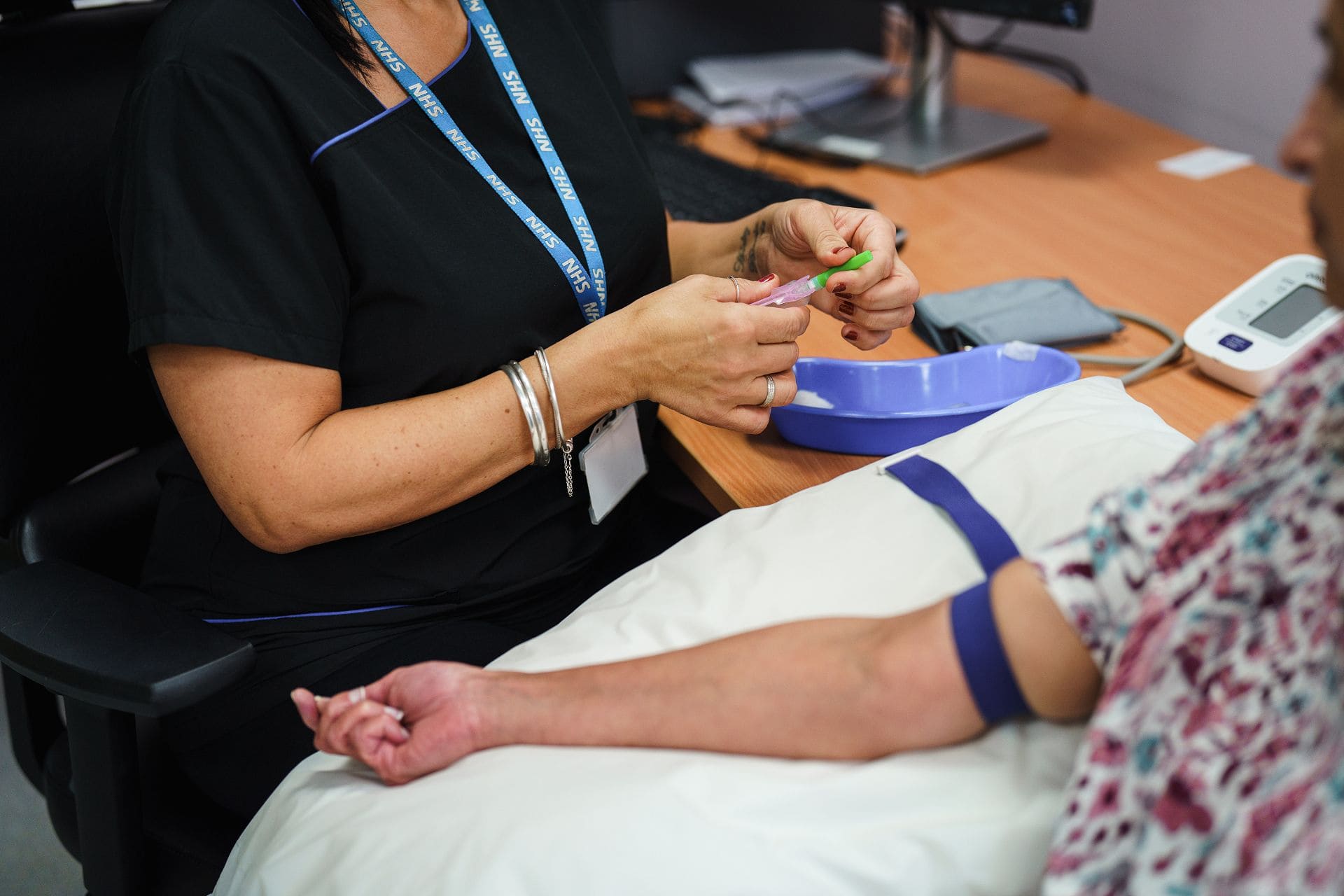 A healthcare worker prepares to draw blood from a patient's arm, who is seated with a tourniquet applied, in a medical office setting.