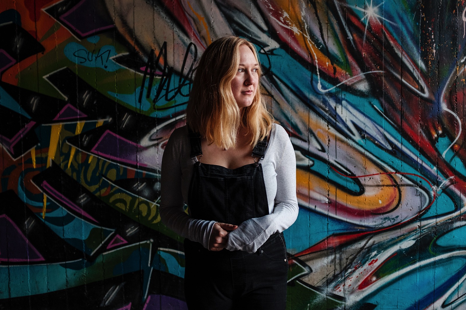 woman in dungarees stood in front of graffiti wall