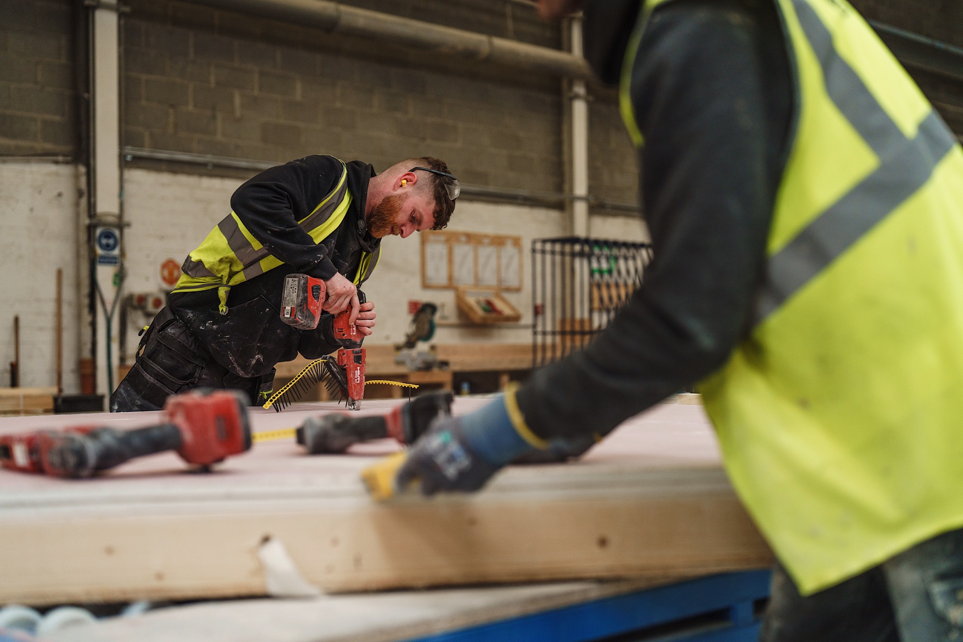 Two workers in safety vests handle drills and wood in a workshop. One focuses on drilling while the other prepares the surface. Industrial tools and materials are visible around them.
