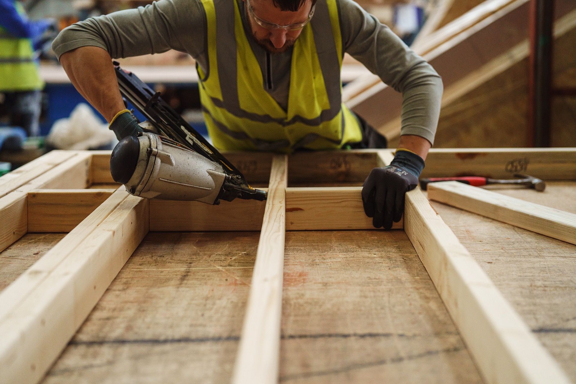 A construction worker in a high-visibility vest uses a nail gun to secure wooden beams on a workbench.