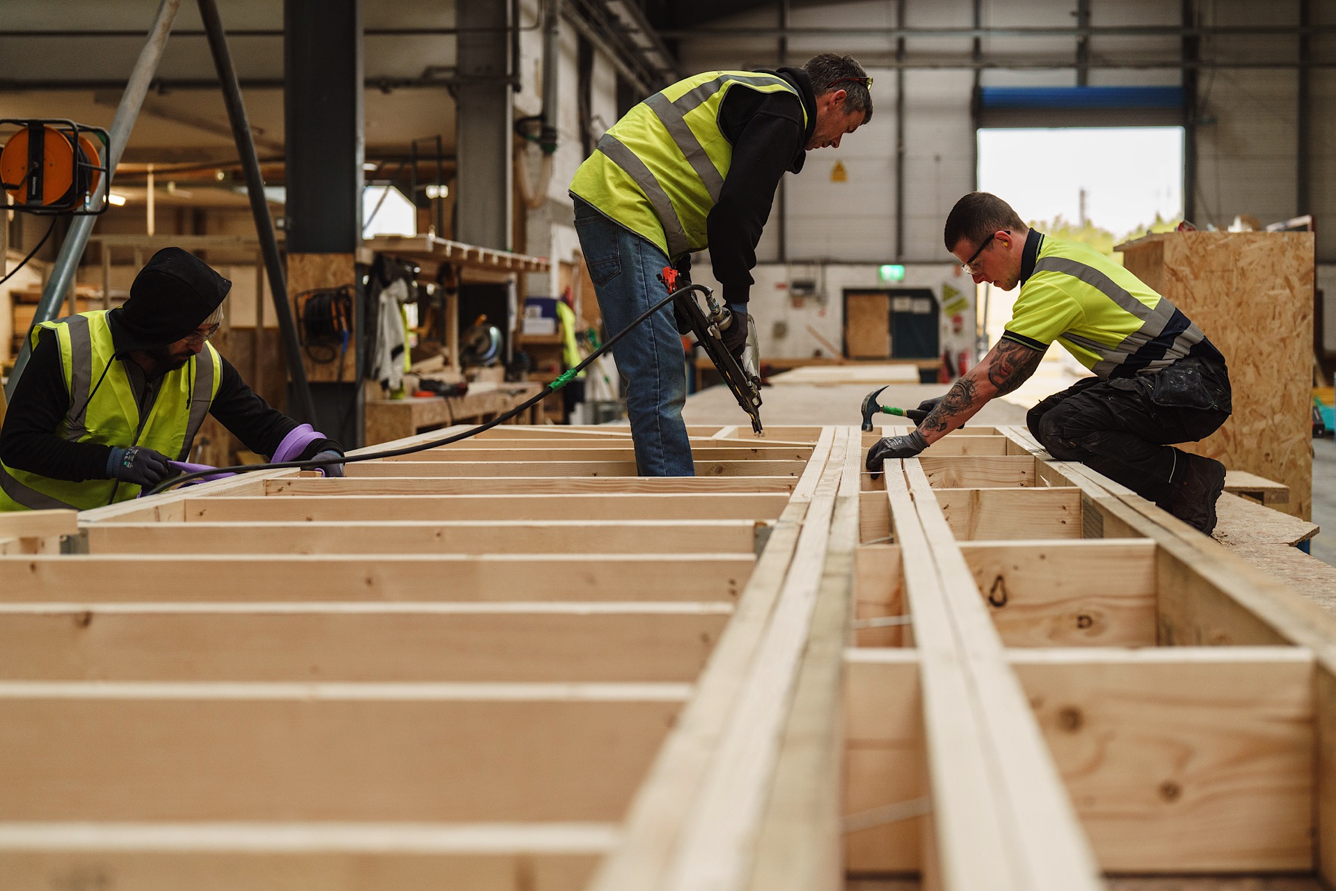 Three workers in safety vests assemble wooden frames inside a warehouse, using tools and protective gear.