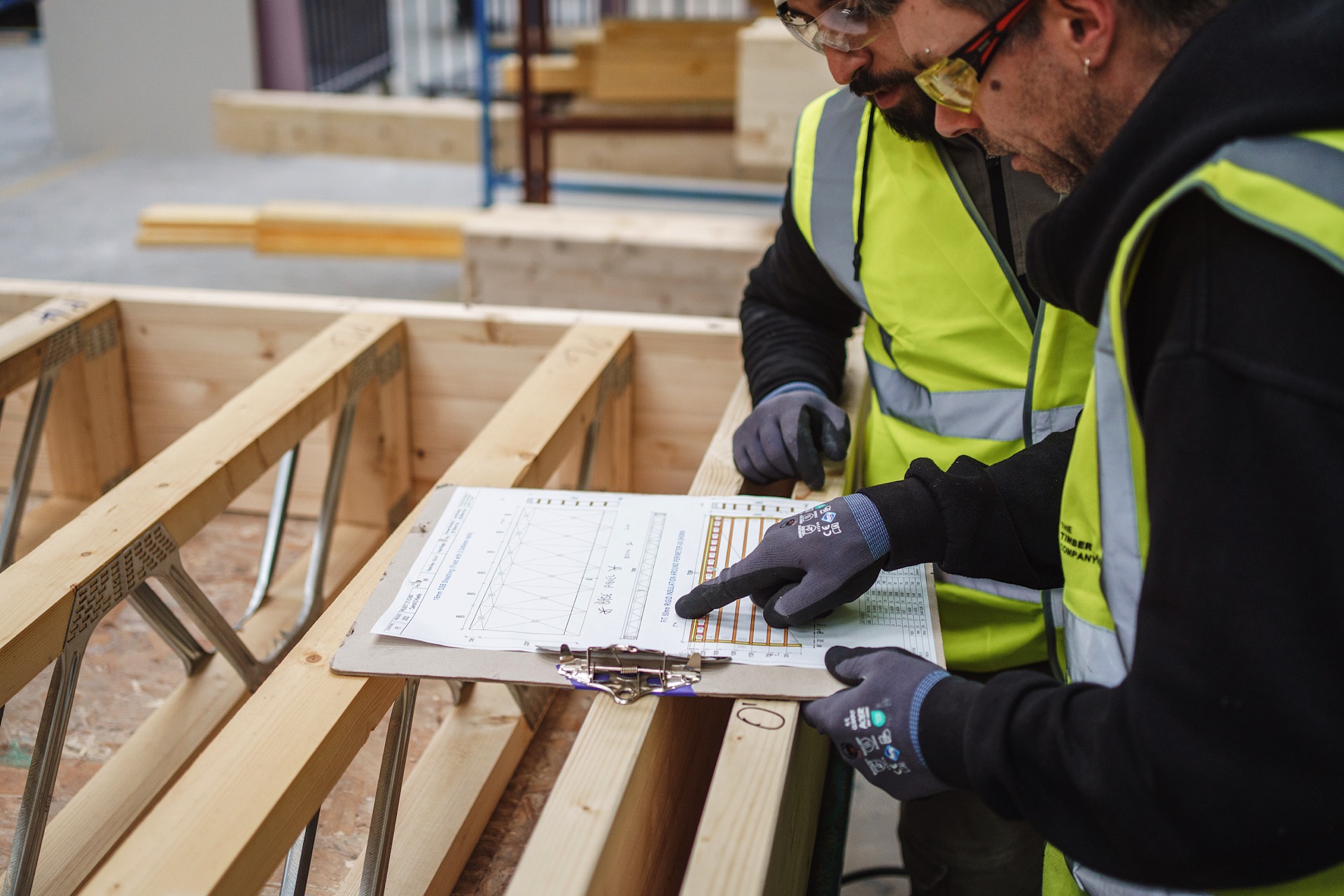 Two people wearing safety vests and gloves review a document on a clipboard over wooden construction beams.