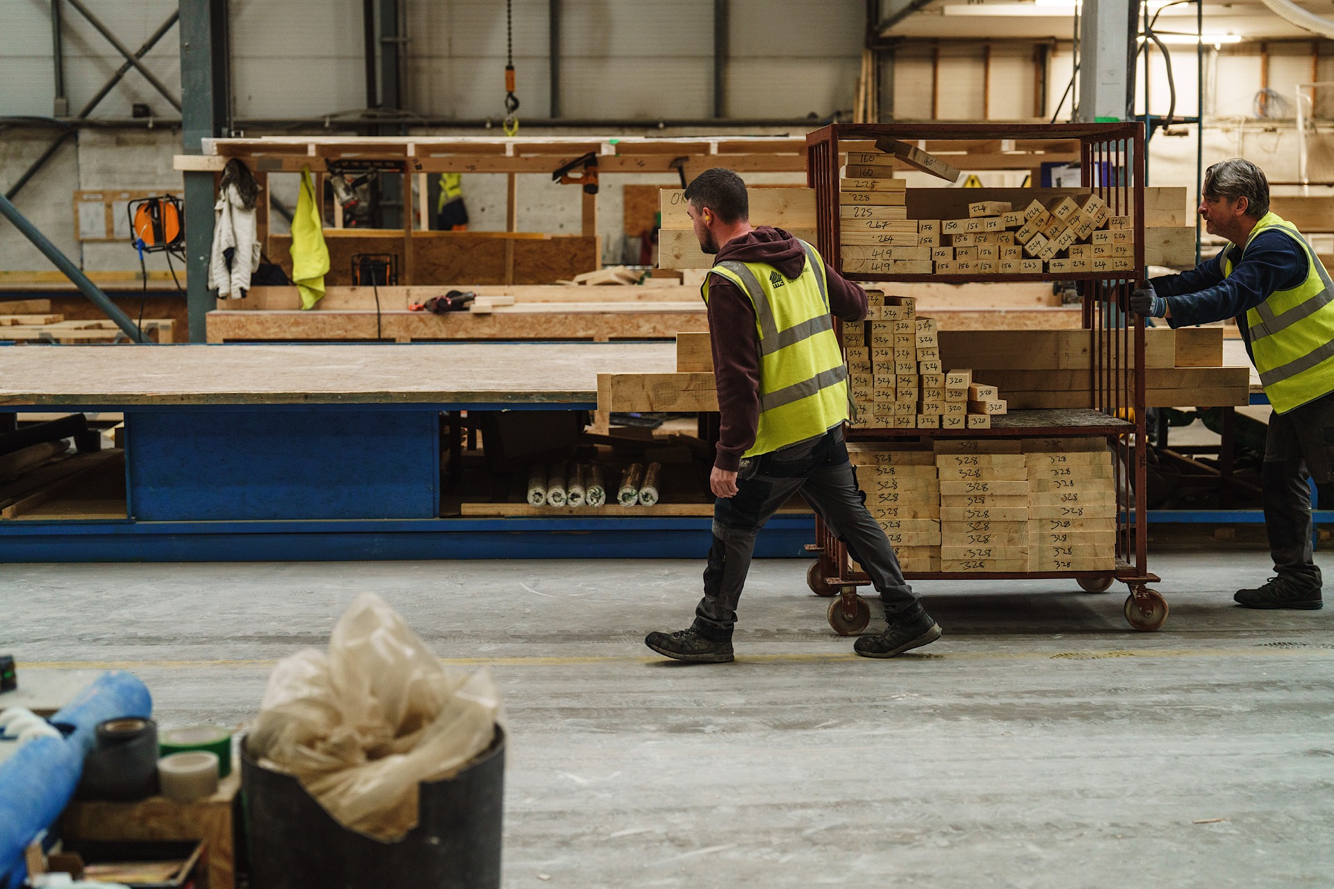 Two workers in a warehouse, wearing reflective vests, push a trolley loaded with materials. Wood and tools are scattered in the background.