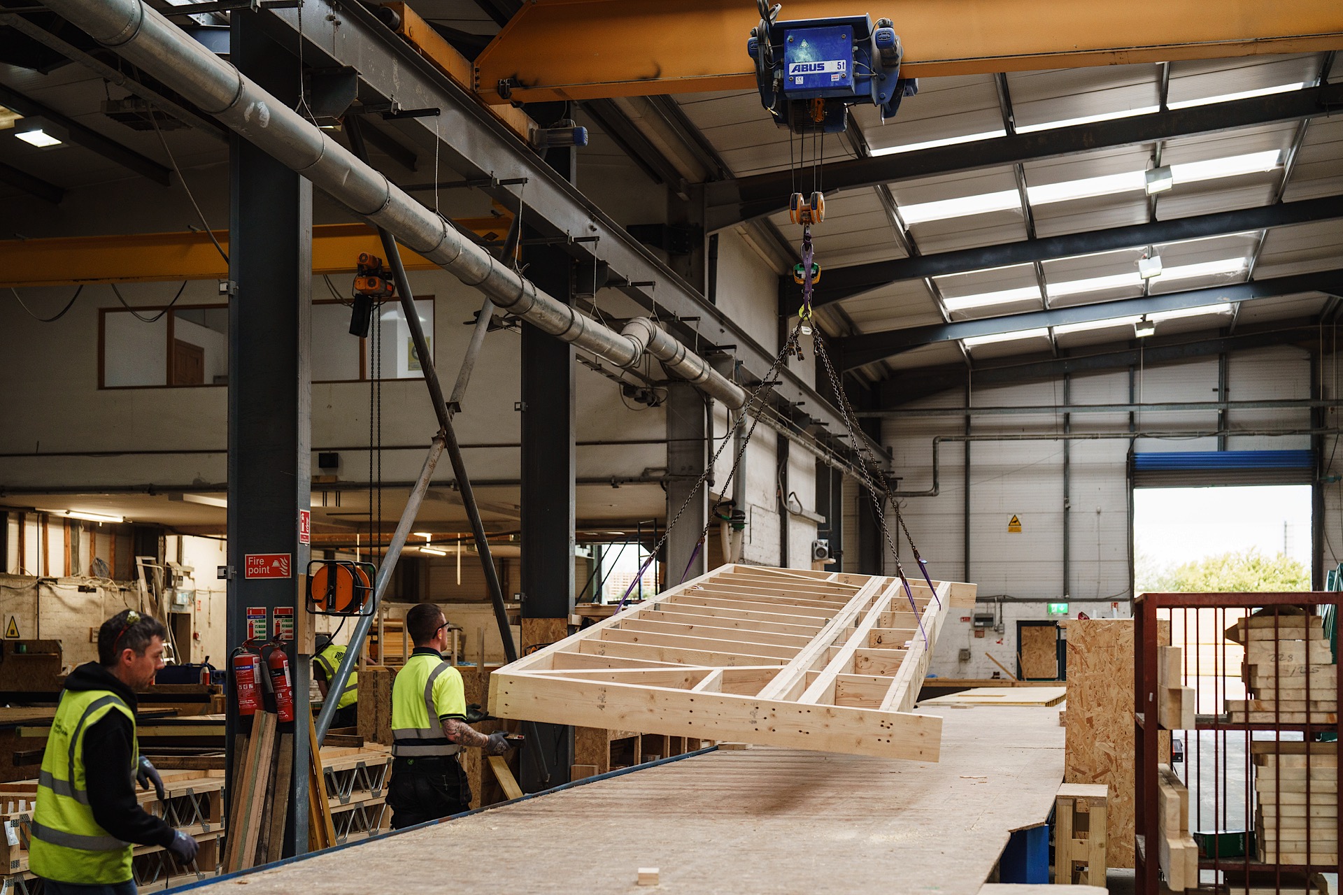 Workers in a factory guide a wooden frame being moved by an overhead crane. The facility has high ceilings and industrial equipment.