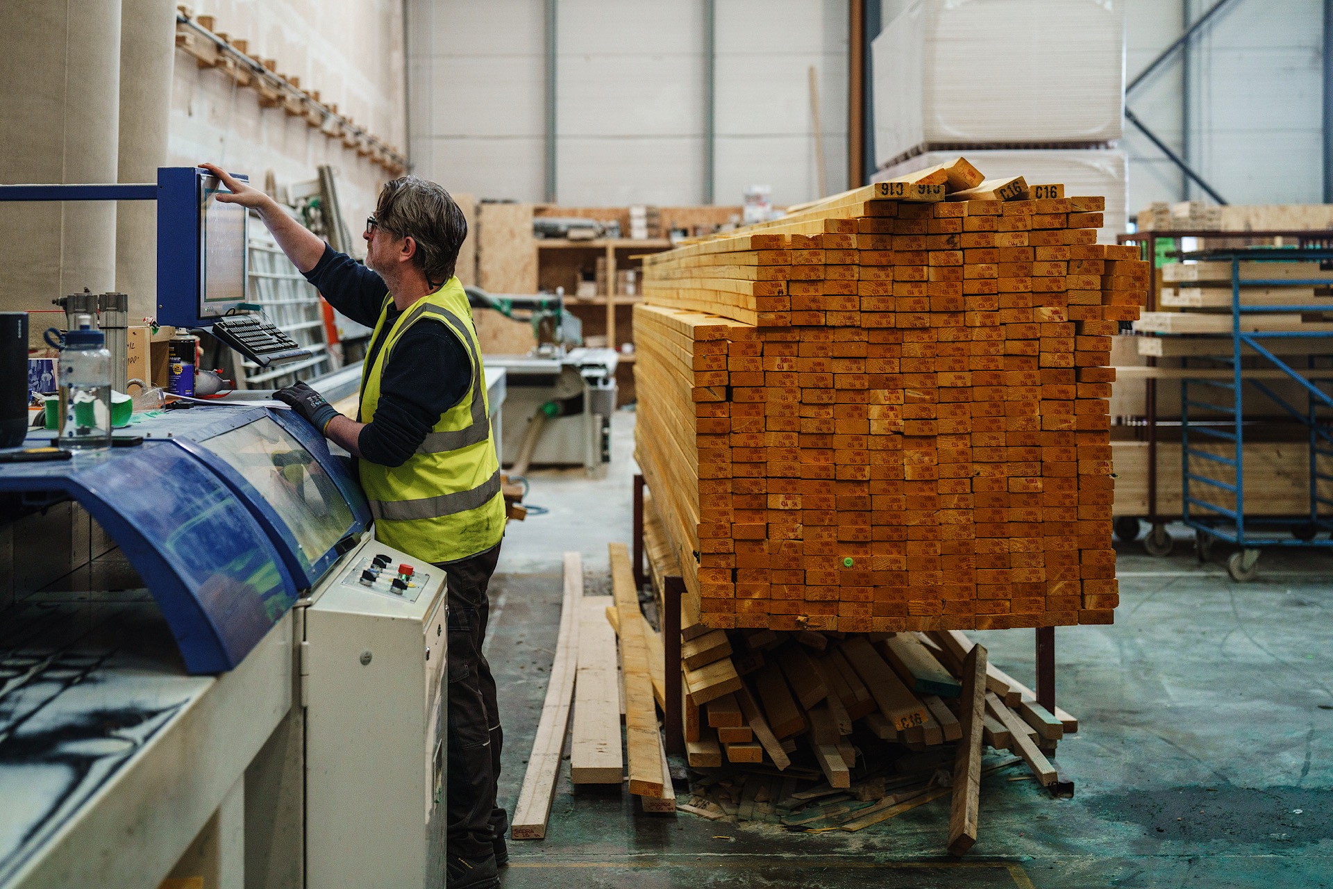 A worker in a high-visibility vest operates machinery in a lumber processing facility, with a large stack of wooden beams visible in the background.