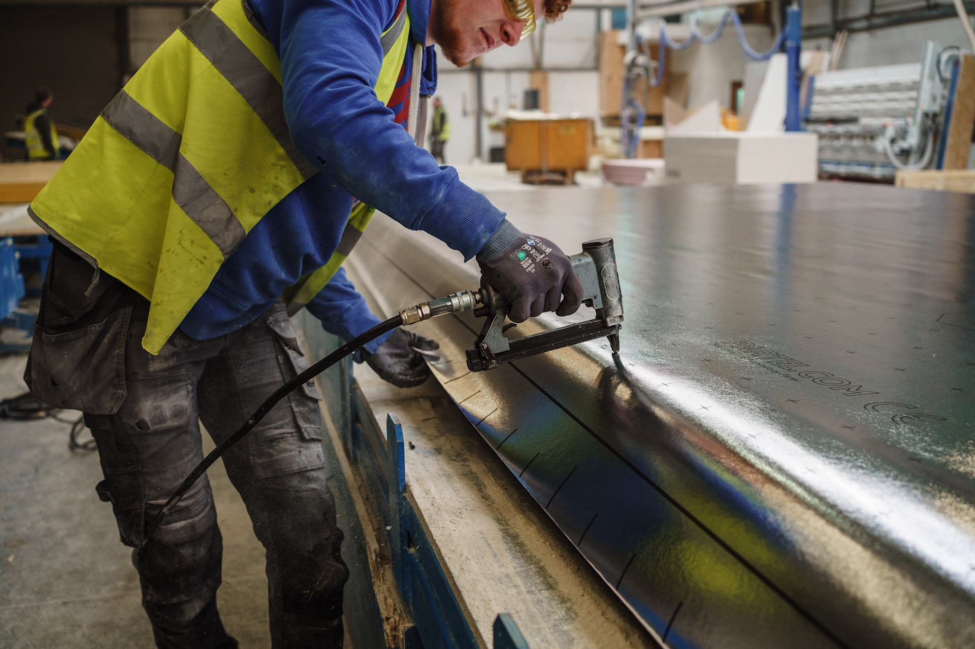 Worker in high-visibility vest and gloves uses a nail gun on a metal sheet in a factory environment.