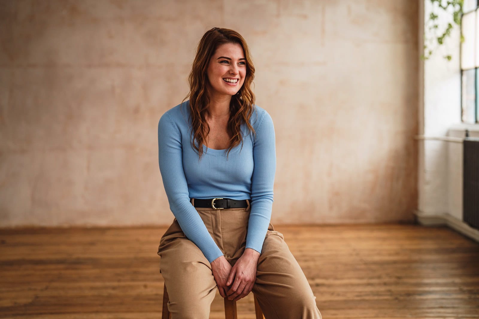 Portrait of a woman wearing a light blue top and khaki pants sat on a stool laughing