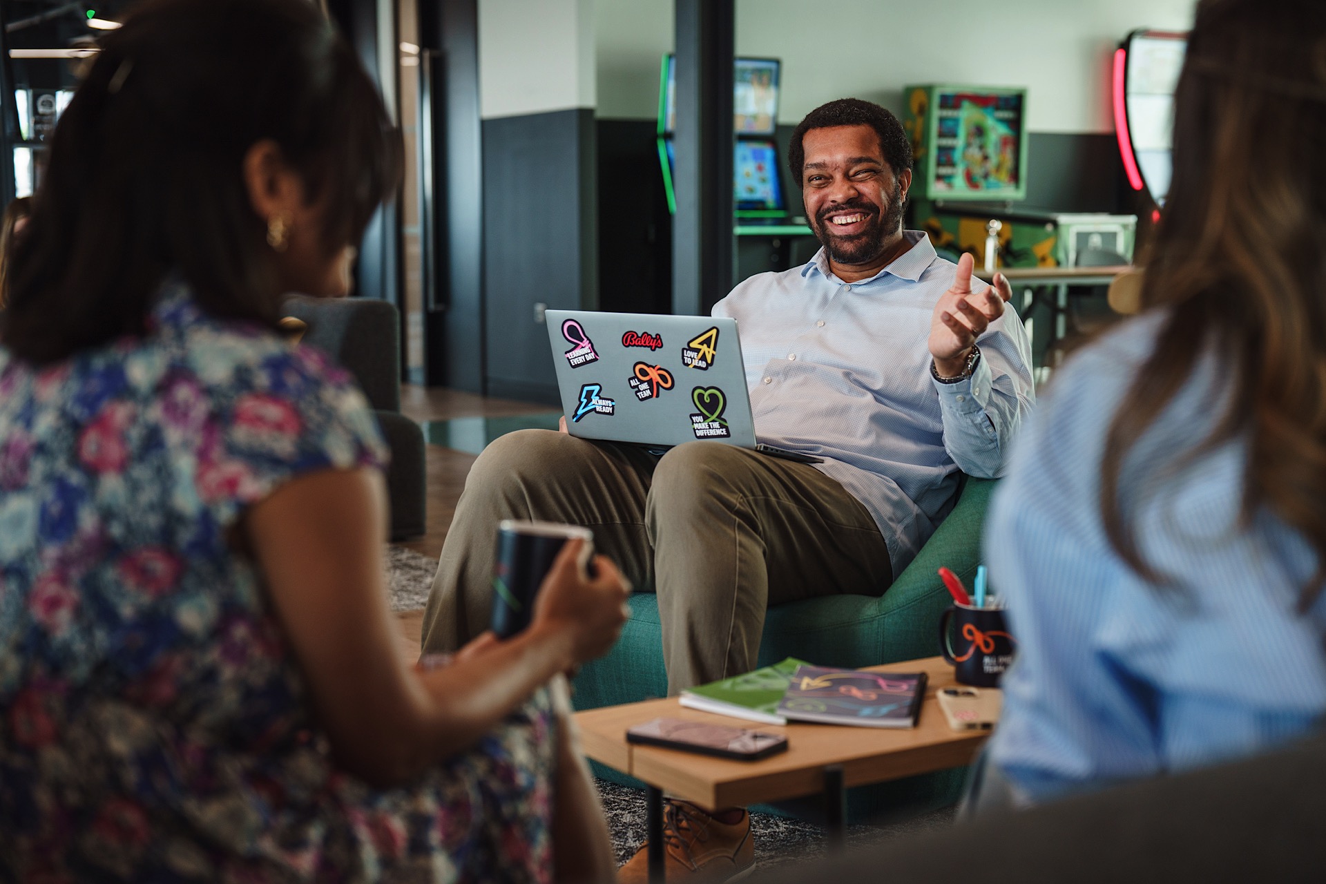 Man sitting on a couch with a laptop covered in stickers, talking to two women in an office lounge.