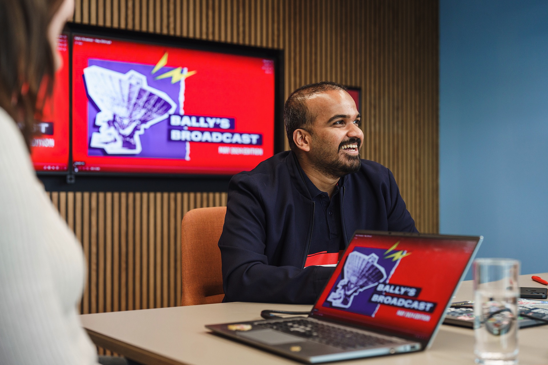 Man seated at a table with a laptop displaying "Bally's Broadcast." Another person sits nearby. A large screen in the background shows the same logo and text.