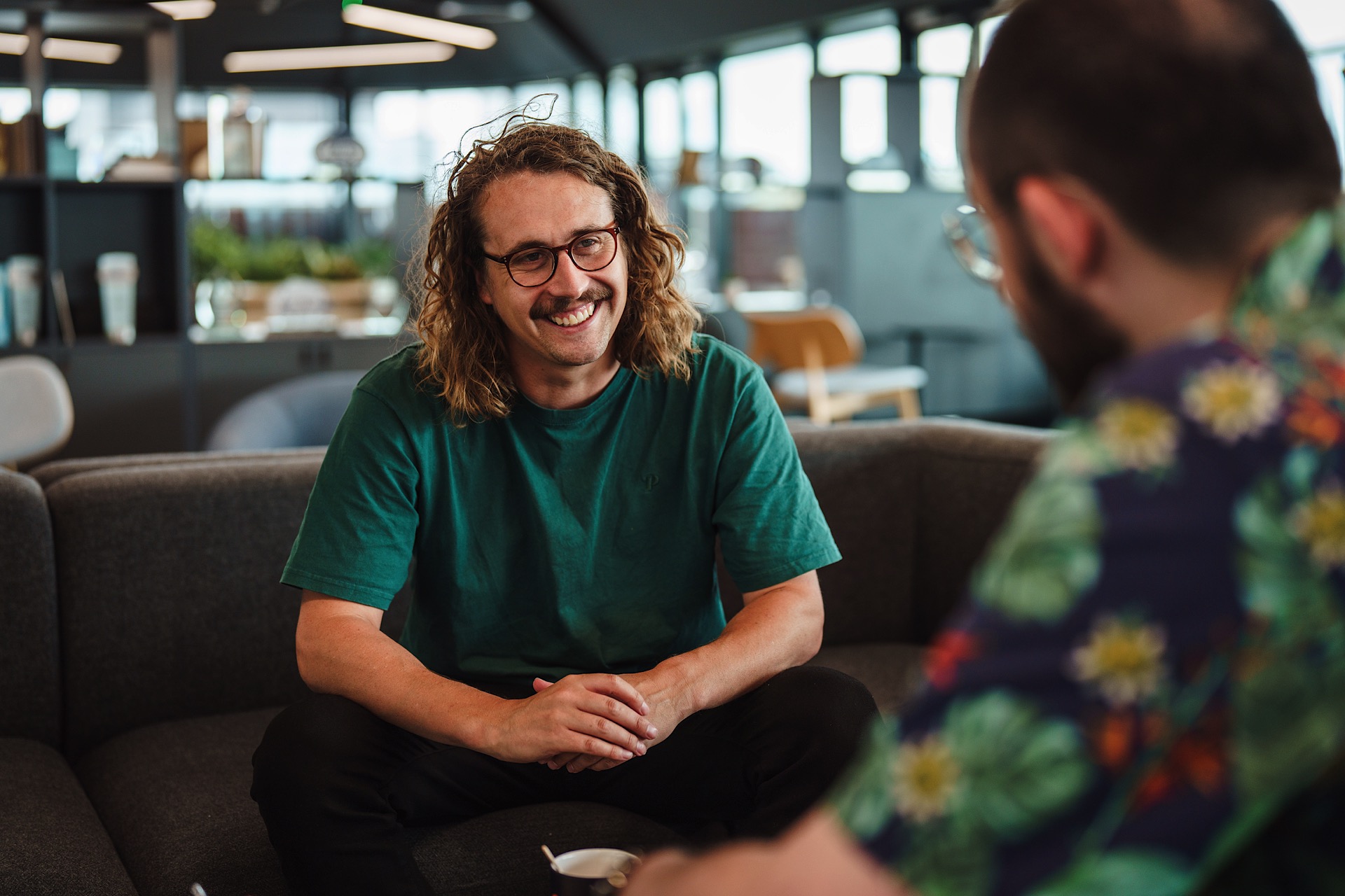 Two men sitting on a couch in a modern office, engaged in conversation. The man facing the camera has long hair and glasses, wearing a green shirt.