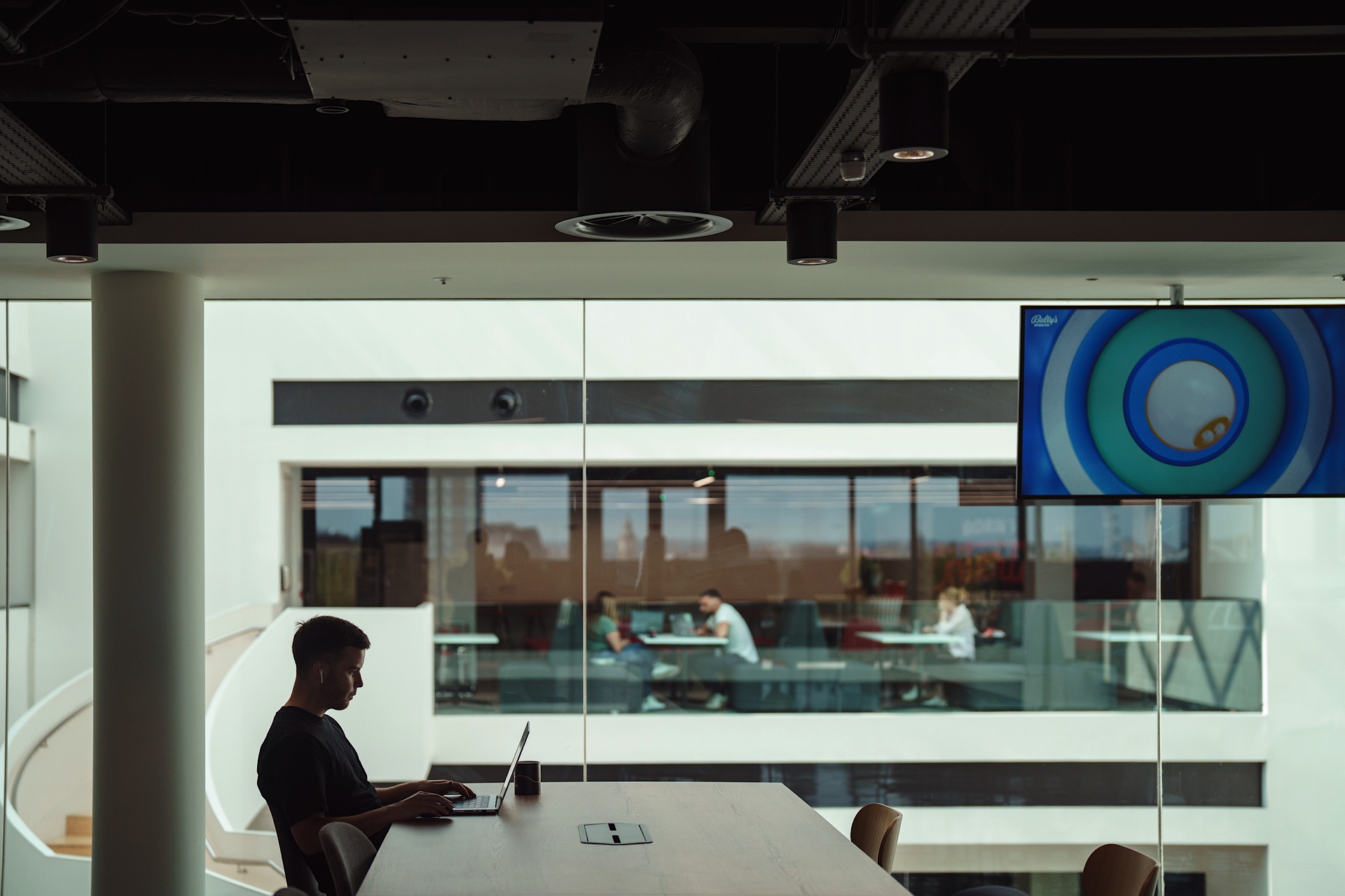 A person working on a laptop at a large table in an office with glass walls. Another building and a wall-mounted TV displaying an animated image are visible in the background.