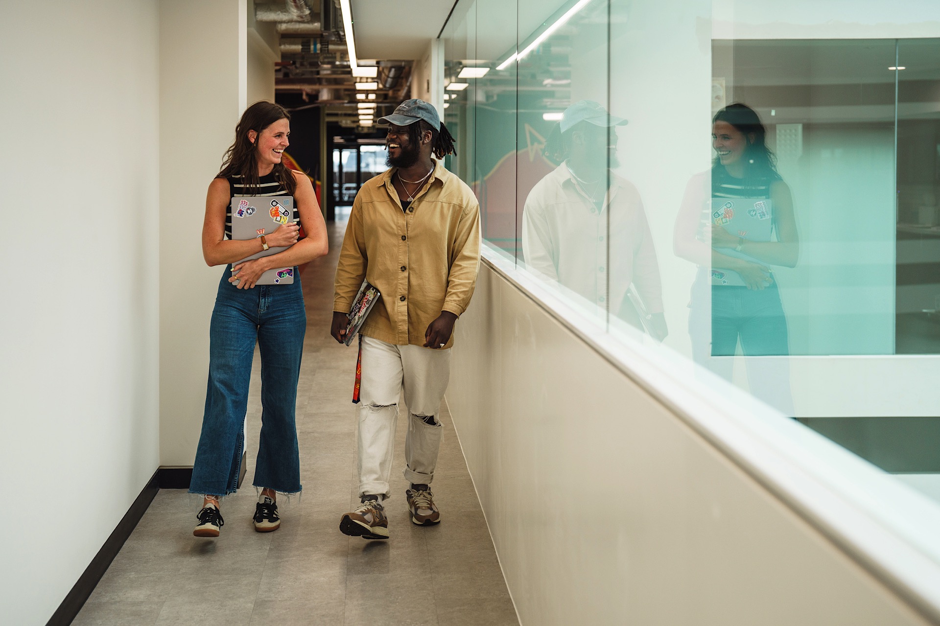 Two people walk down a hallway, smiling and holding books, with a glass wall on the right.