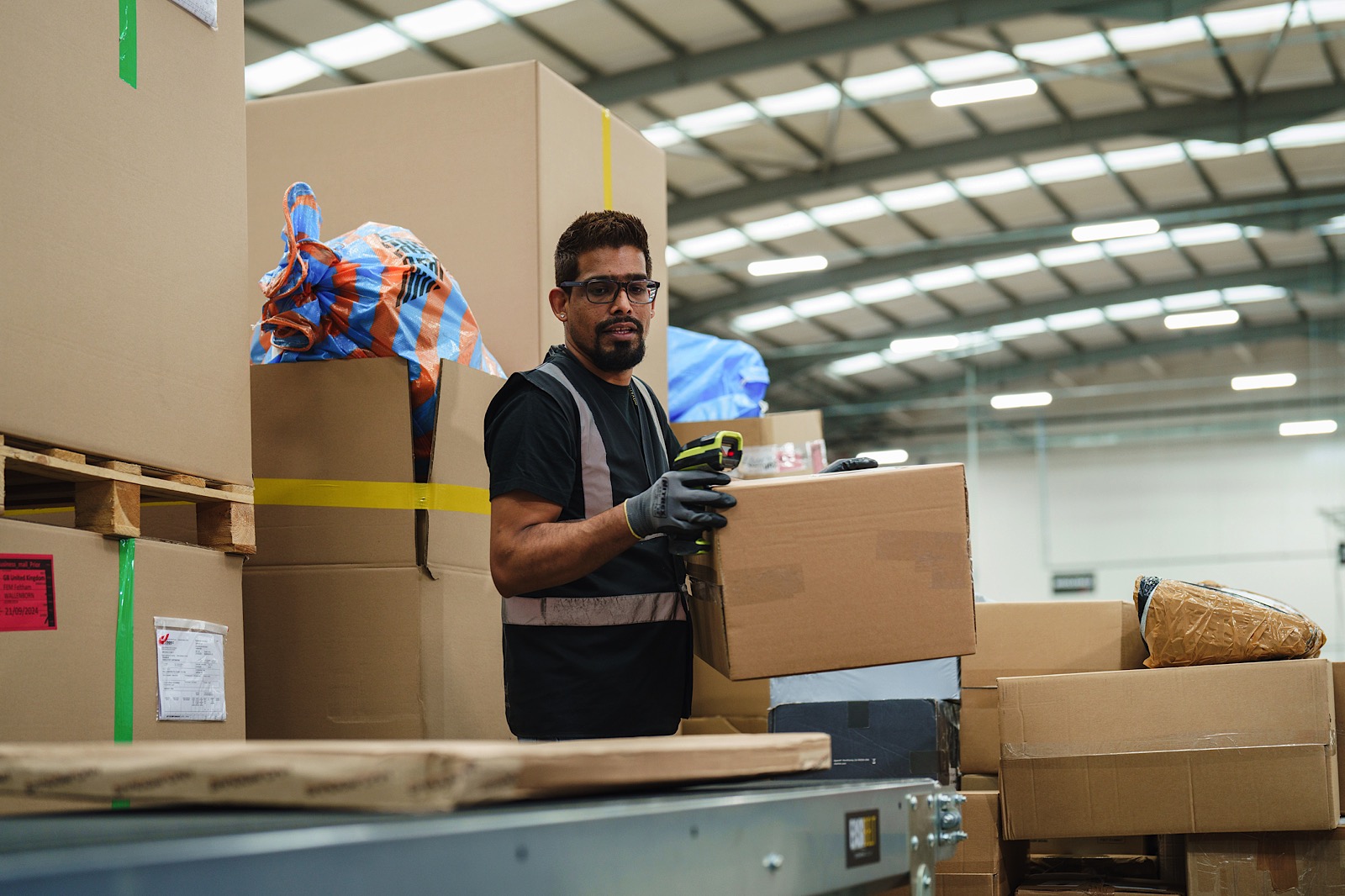 A warehouse worker wearing gloves and a reflective vest holds a cardboard box among stacks of packages inside a large warehouse.