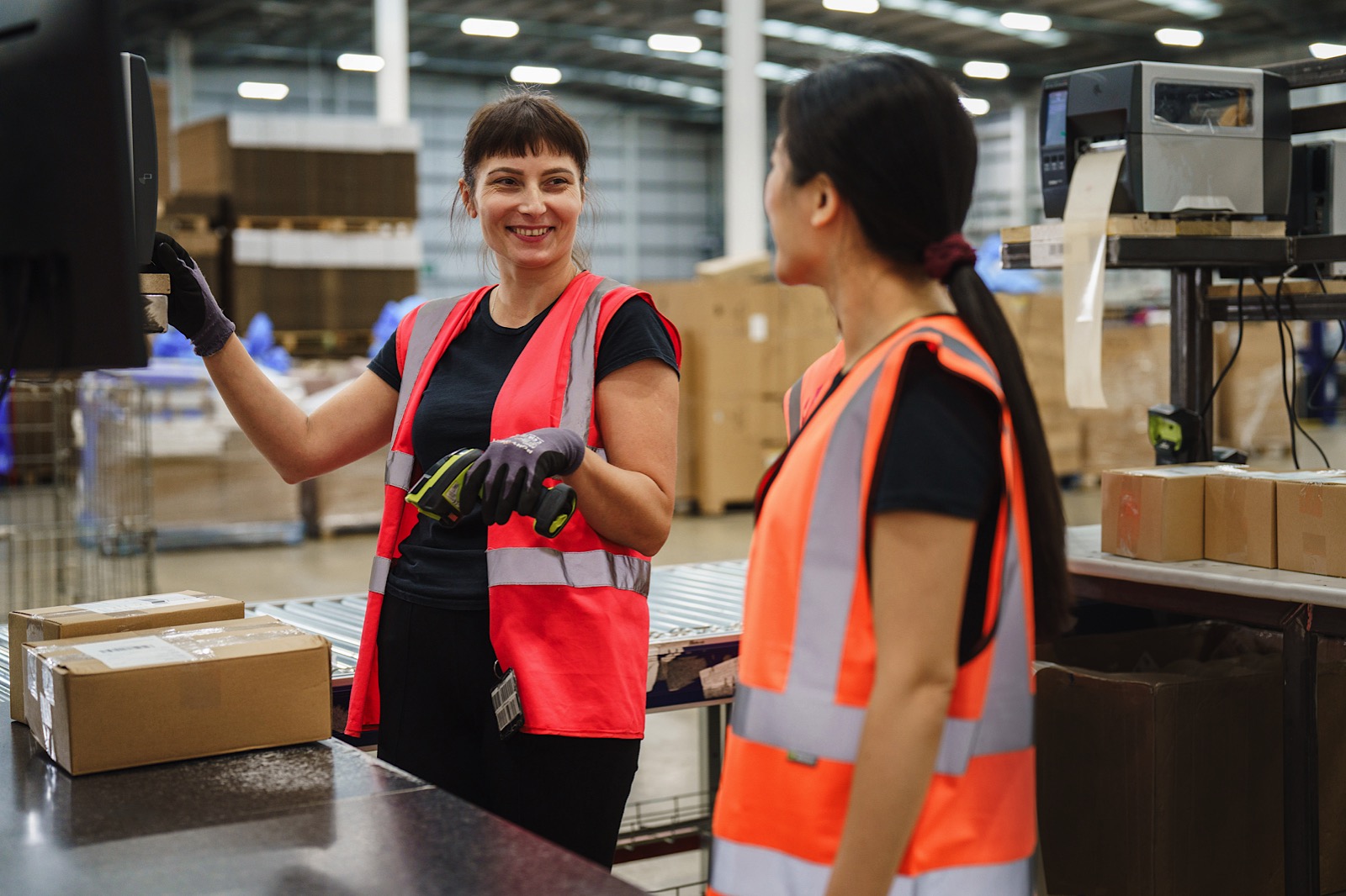 Two warehouse workers wearing orange safety vests and gloves stand by a workstation with packages, talking and smiling in a large storage facility.