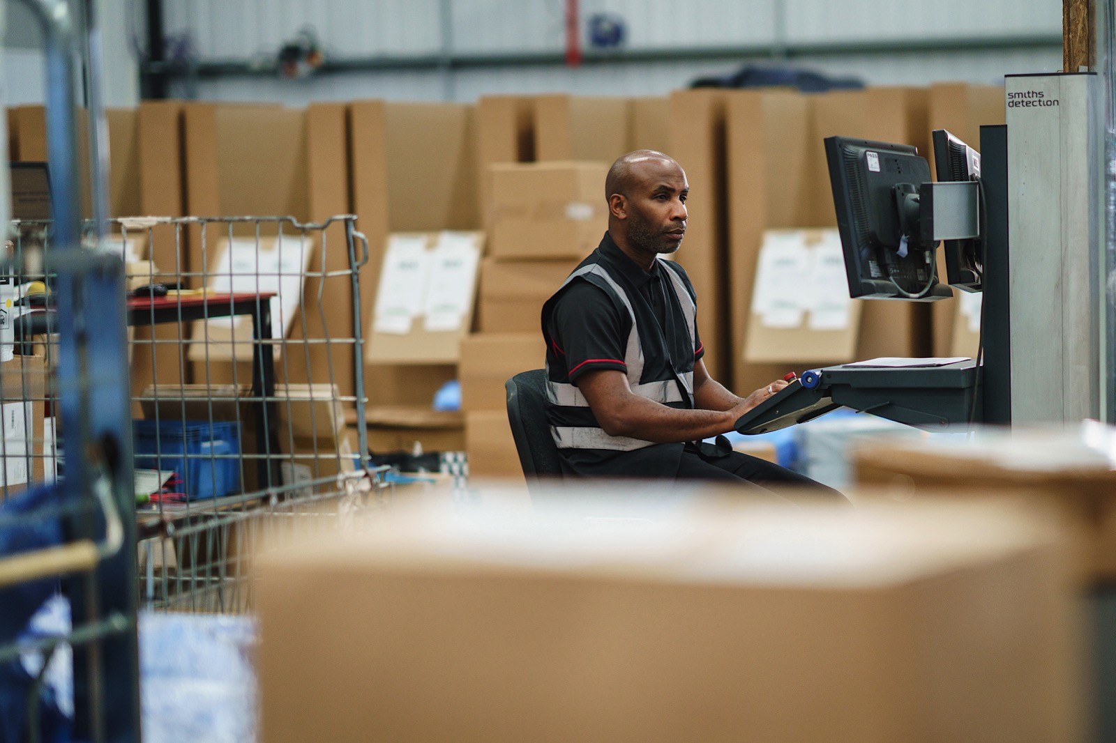 A man sits at a desk using a computer in a warehouse, surrounded by large cardboard boxes and industrial shelving.