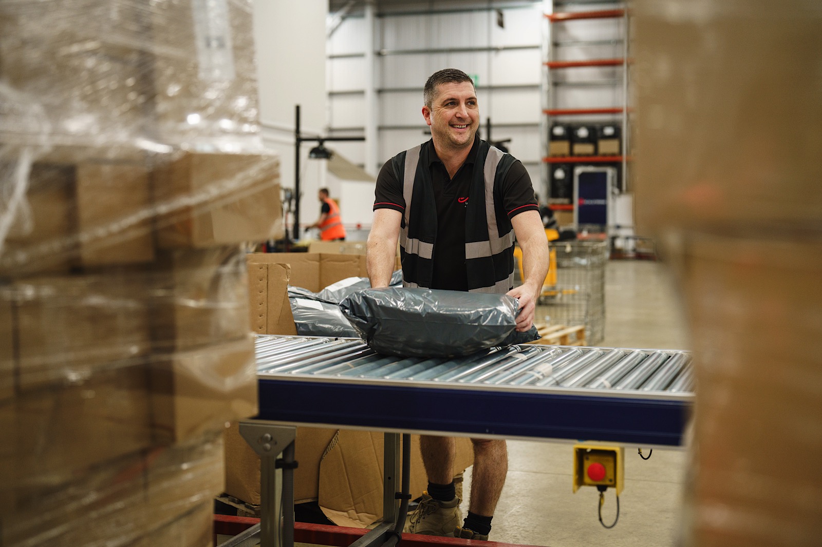 A man wearing a safety vest places a package on a conveyor belt in a warehouse surrounded by cardboard boxes.