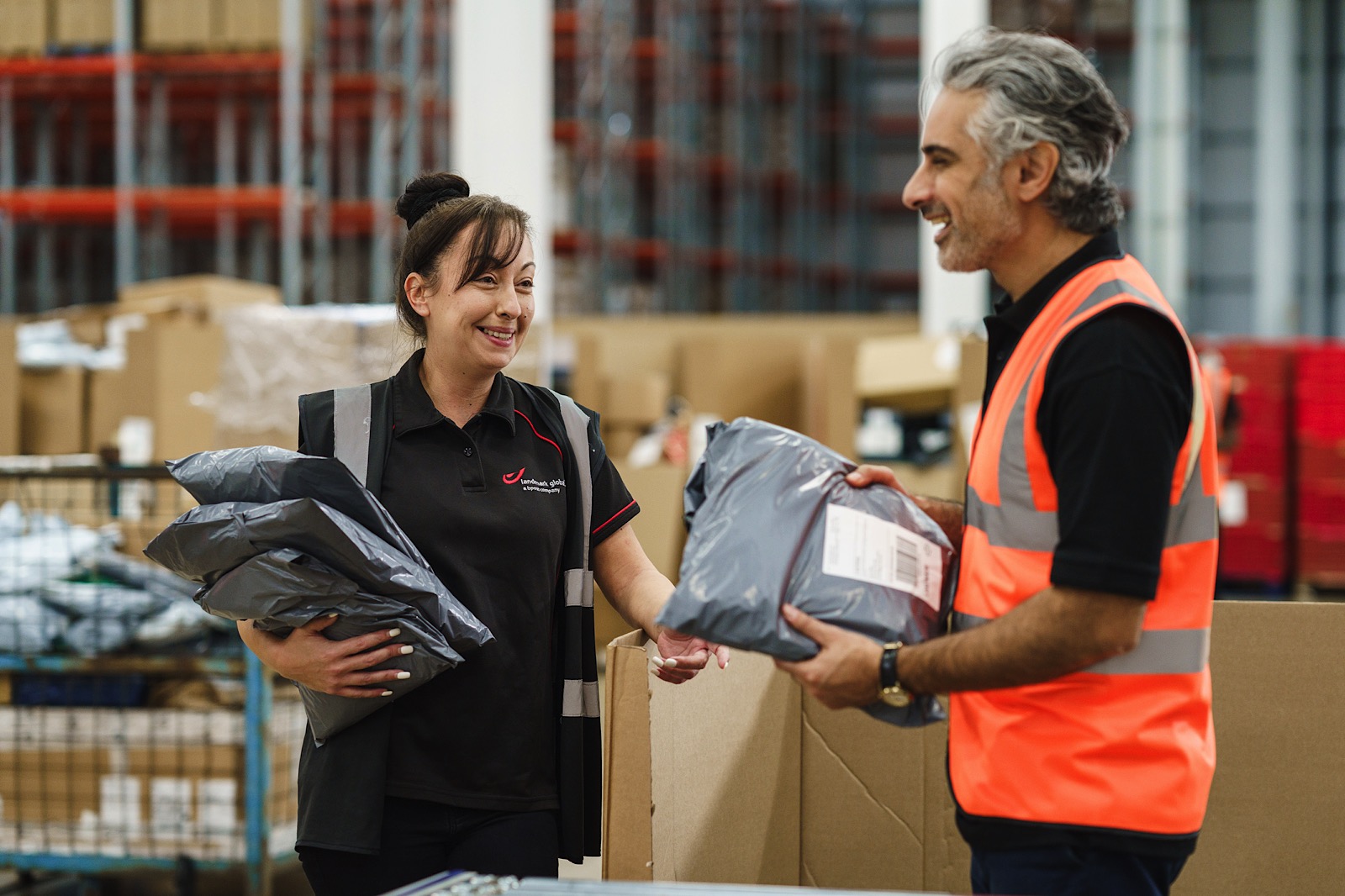 Two warehouse workers, one holding parcels and the other wearing a high-visibility vest, stand and talk in a warehouse surrounded by boxes and shelves.