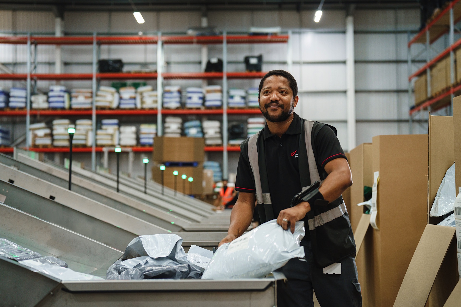 A warehouse worker sorts parcels on a conveyor belt, surrounded by shelves stocked with various items in an industrial setting.