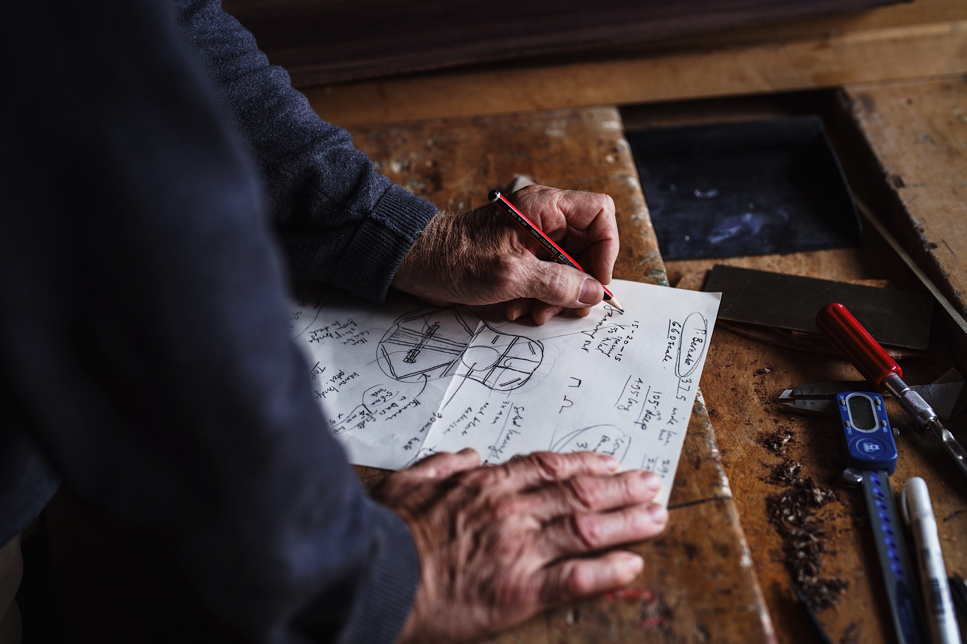 A person sketches a detailed design on paper at a wooden table, surrounded by drafting tools and a red pencil.