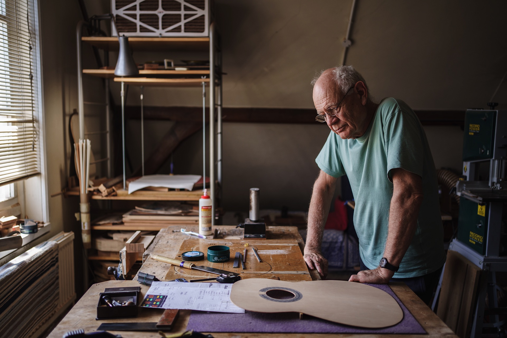 Nick Branwell leaning over a table with guitar-making tools and materials, including a partially constructed guitar, under soft lighting.