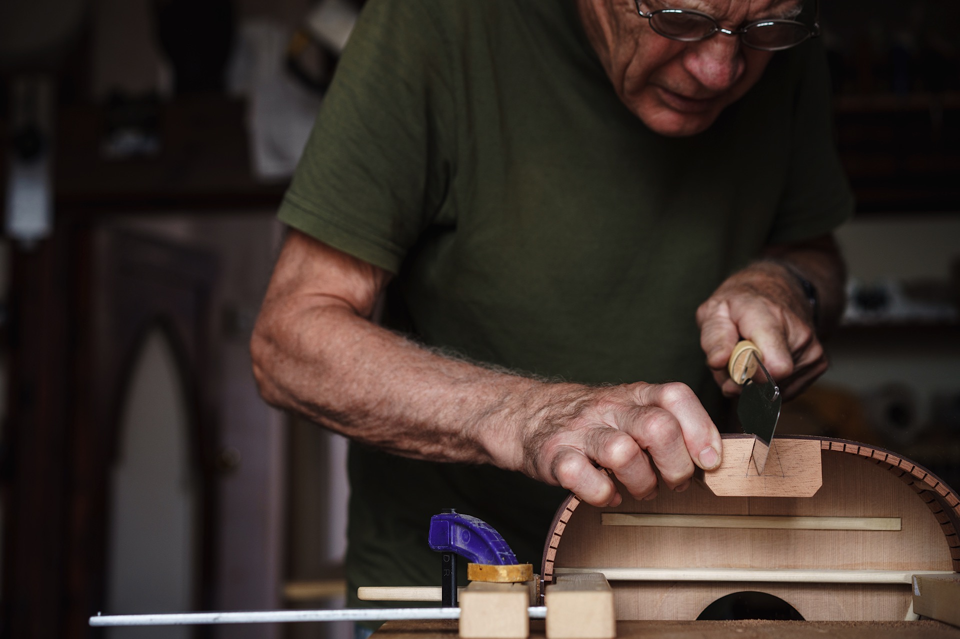 Nick Branwell wearing a green shirt is using a knife to shape a guitar in his workshop, with clamps and tools visible on the table.