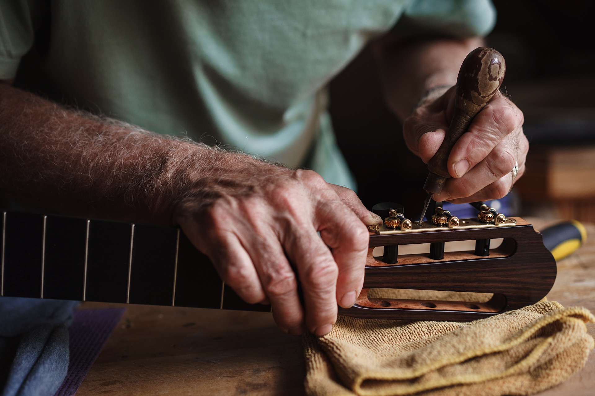 A person adjusting the tuning pegs of a guitar with a tool, seated at a wooden table