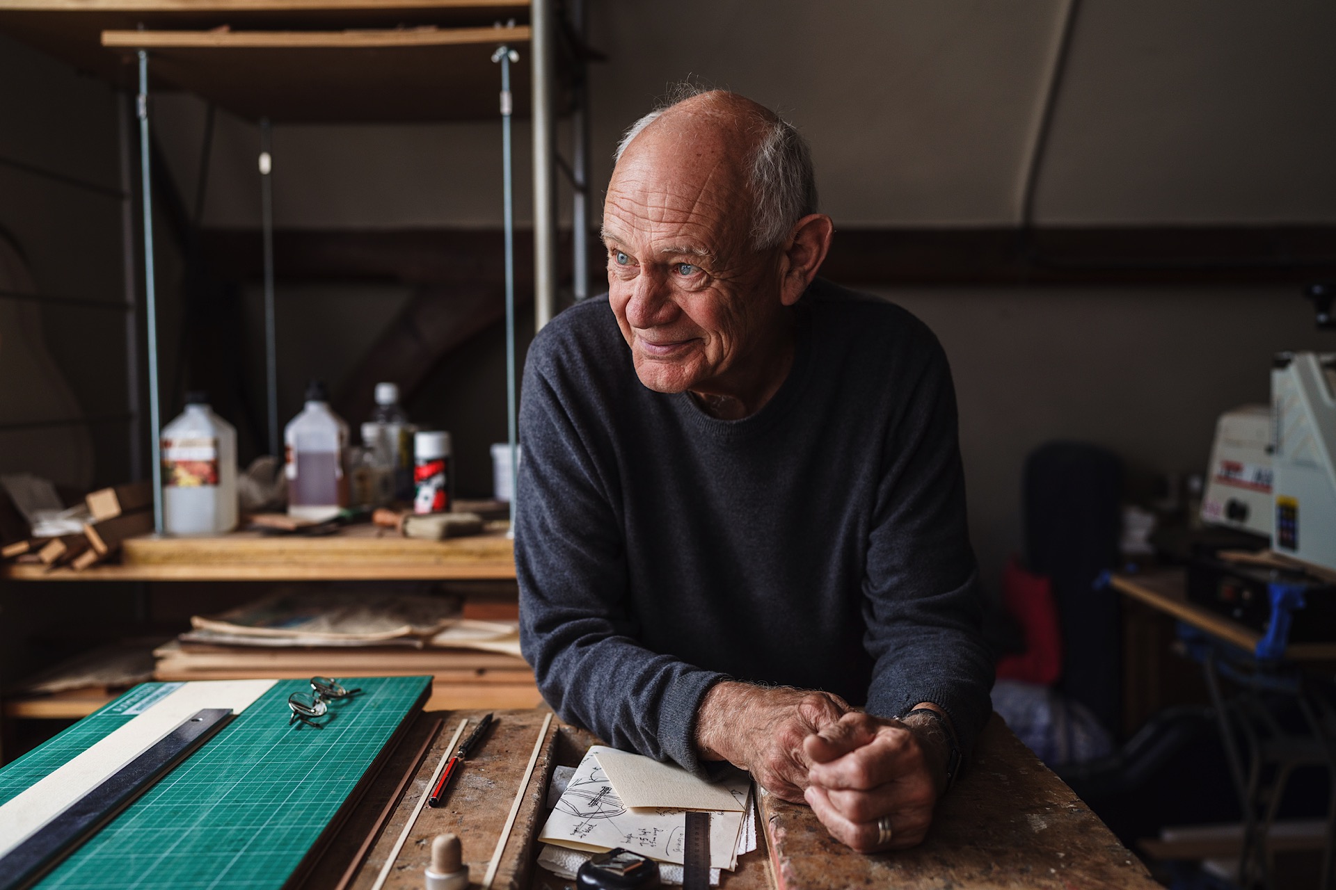 Nick Branwell, Luthier wearing a gray sweater stands leaning on a workshop table with art supplies, looking to the side.
