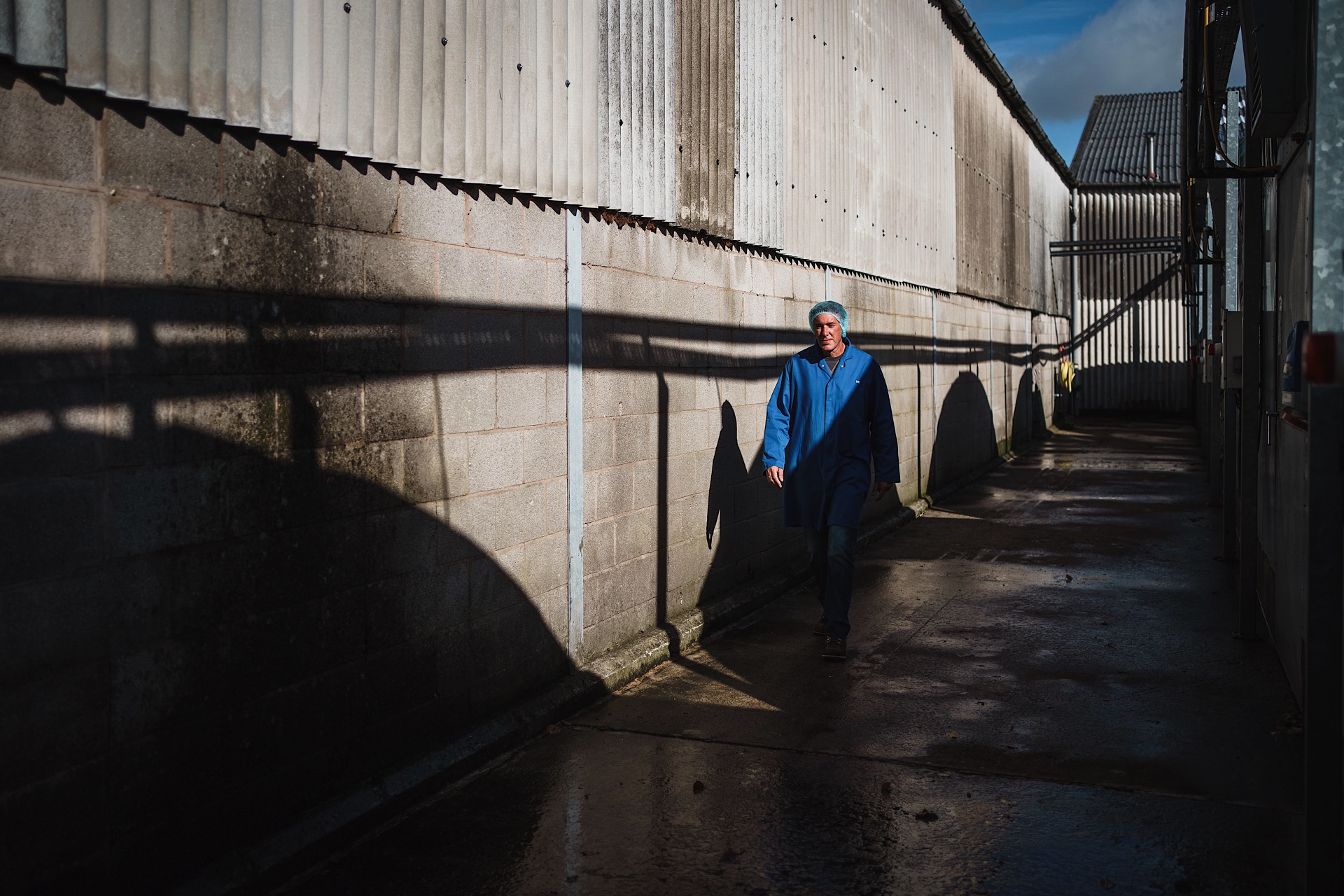 A person in a blue jacket and cap walks down a narrow, sunlit alley between industrial buildings. Shadows are cast on the ground and wall.