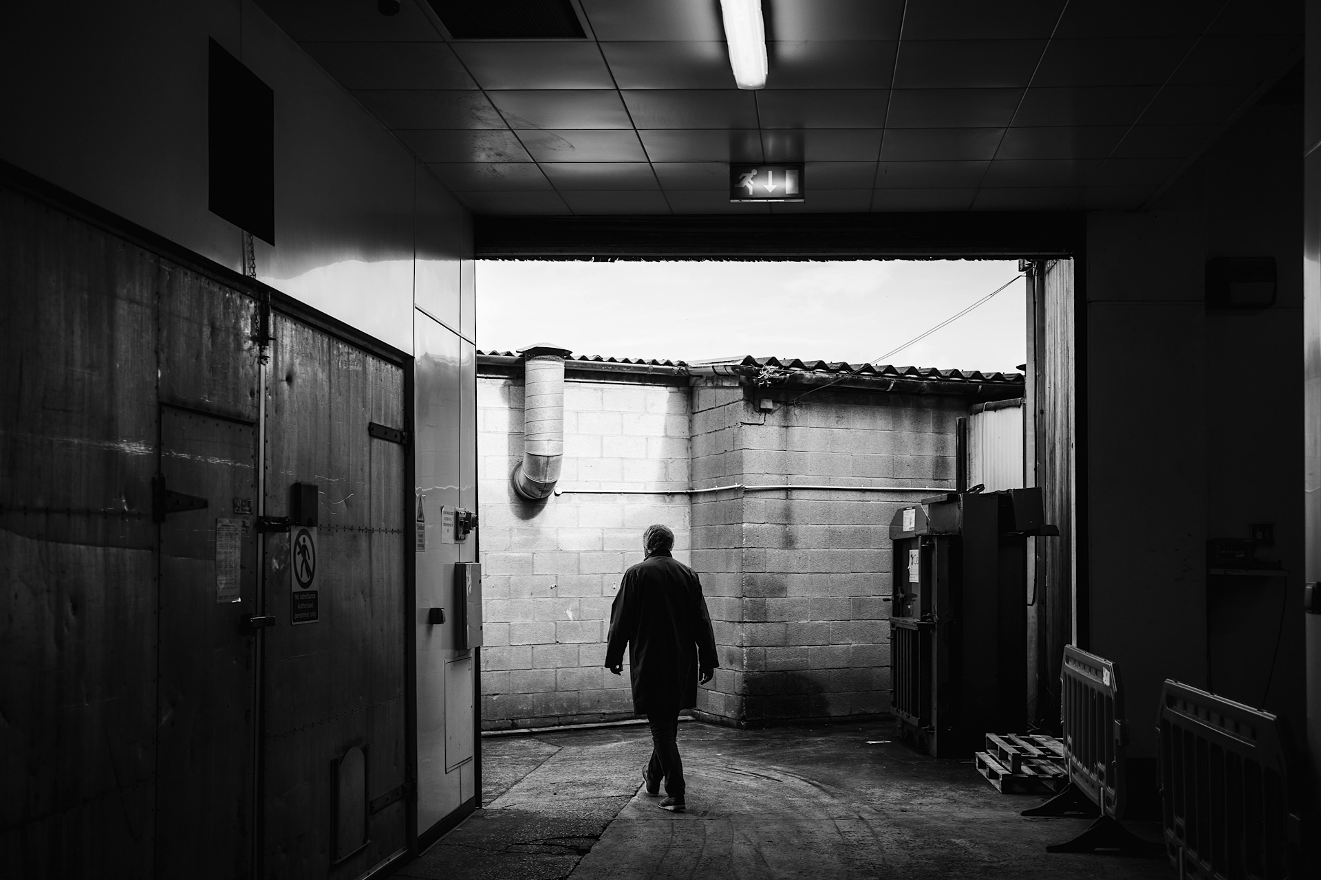 A person walks through an industrial hallway towards an outdoor area, with walls and a roof visible. The scene is in black and white.
